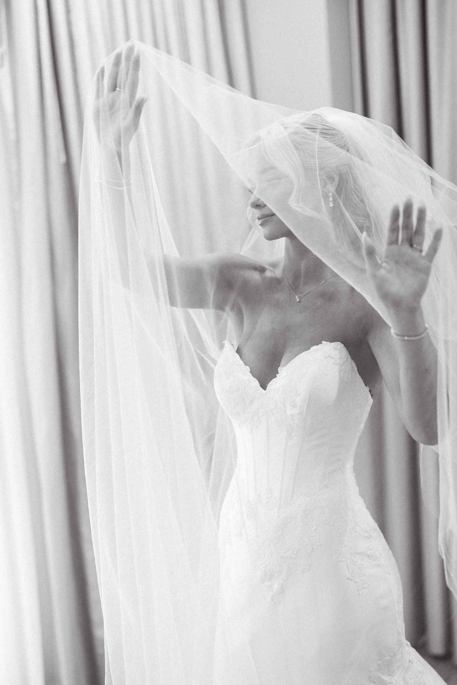 Bride in strapless lace wedding gown lifts flowing veil overhead in elegant black and white bridal portrait.