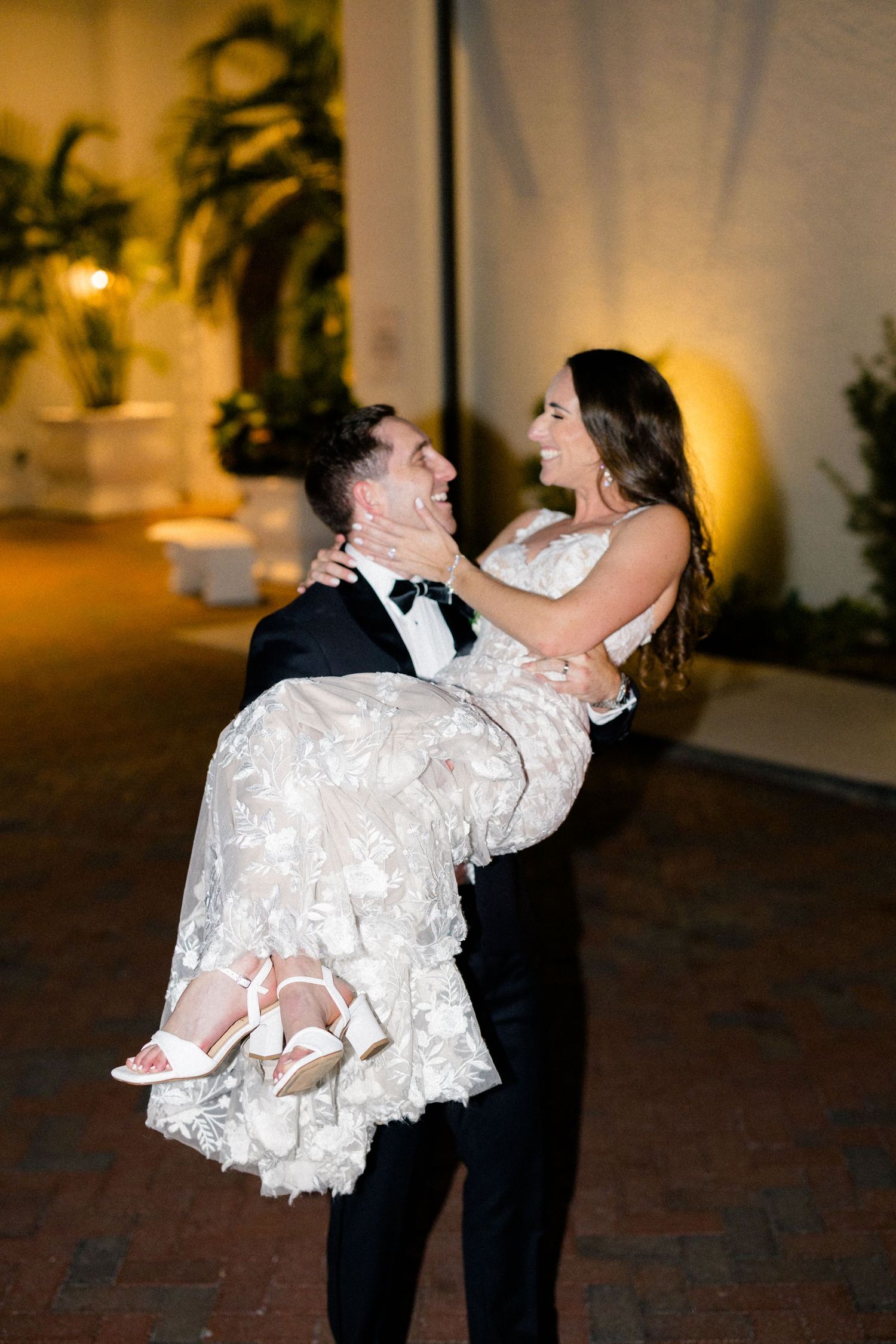 Groom in tuxedo joyfully lifts smiling bride in lace wedding gown outdoors at night.