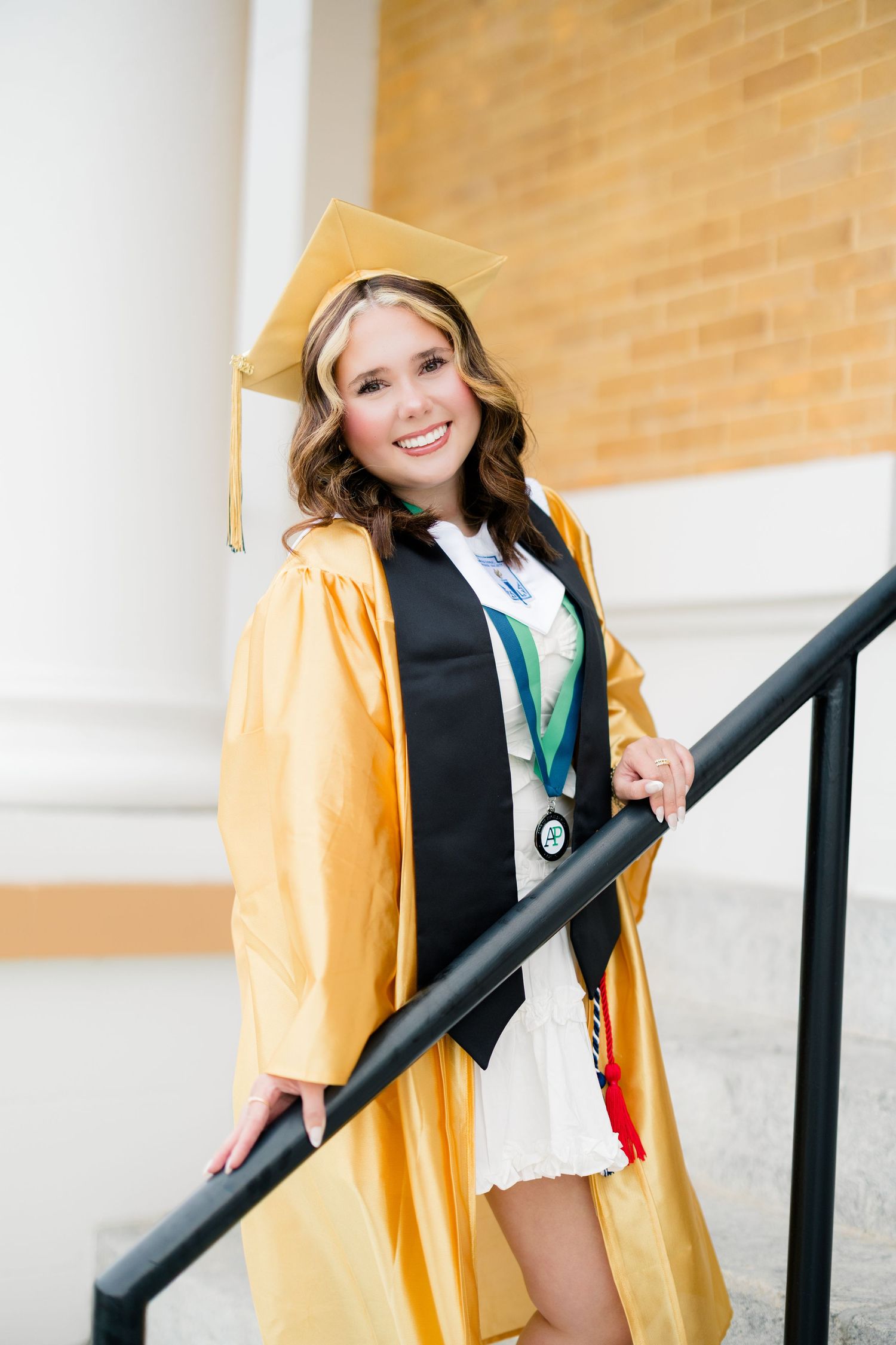 Smiling graduate in gold cap and gown poses on staircase with medal and honor stole.