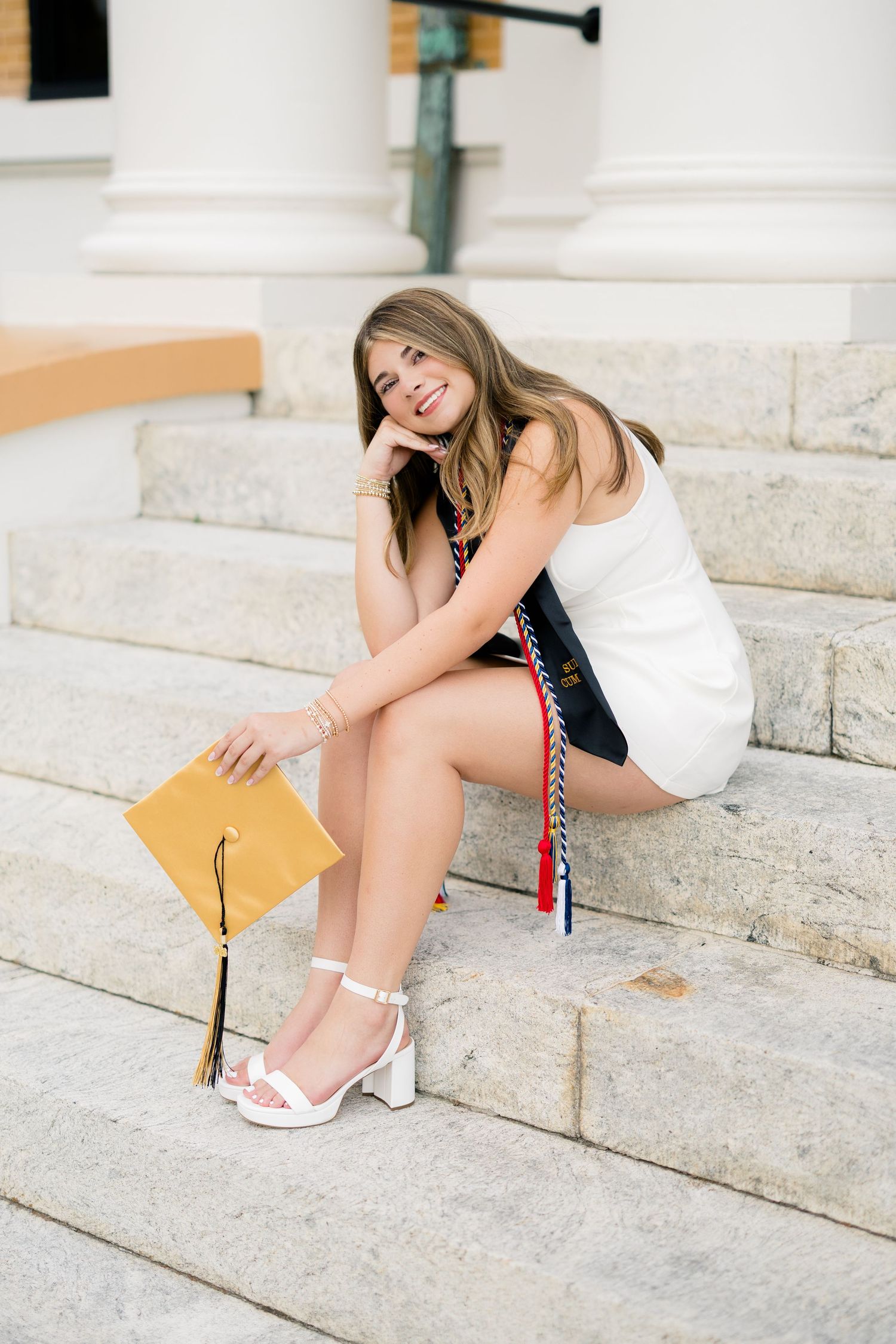 Smiling graduate in white dress sitting on stone steps holding gold mortarboard cap with tassel and colorful honor cords.