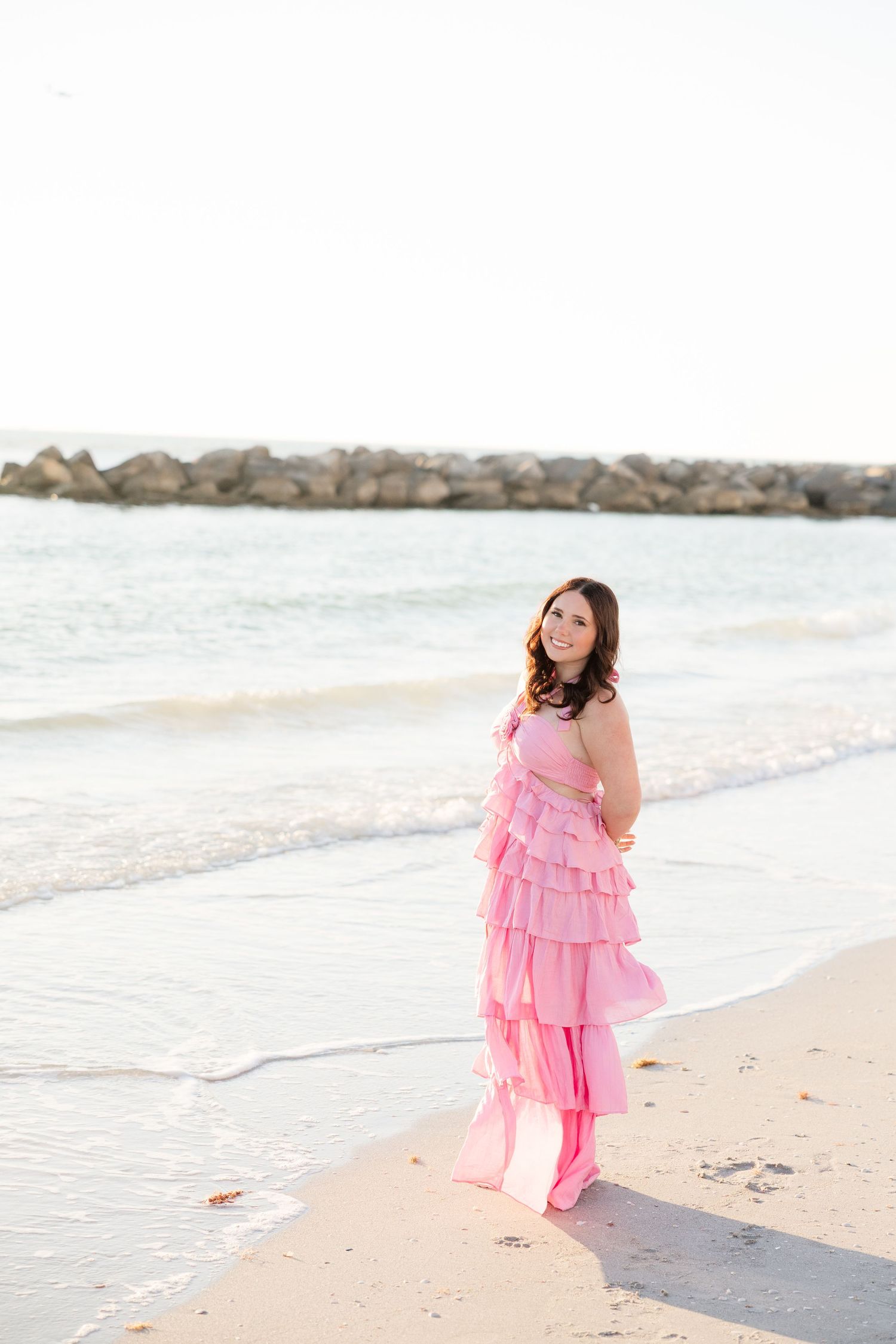 Woman in pink ruffled dress and boots posing on sandy beach with ocean waves and rock jetty behind her.