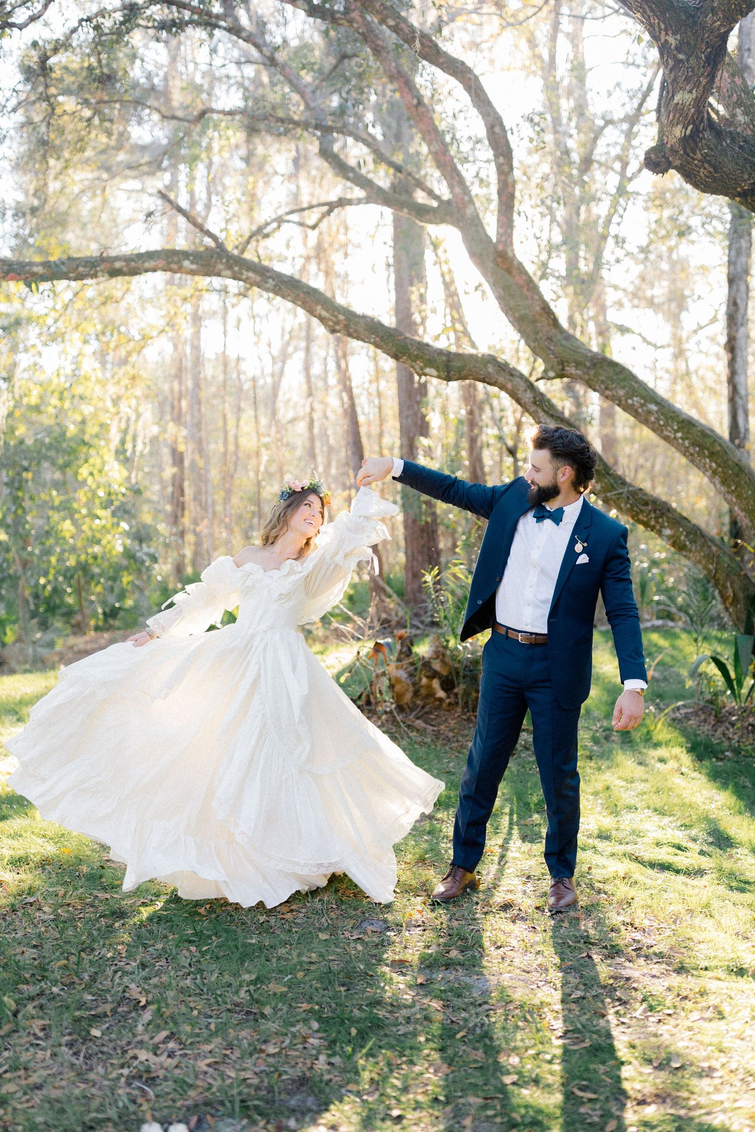 Bride in white gown twirling with groom in navy suit outdoors in sunlit forest setting.