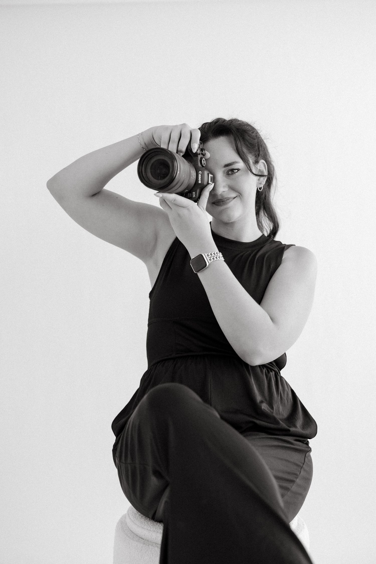Black and white portrait of a smiling woman in black outfit holding a DSLR camera while seated against white background.