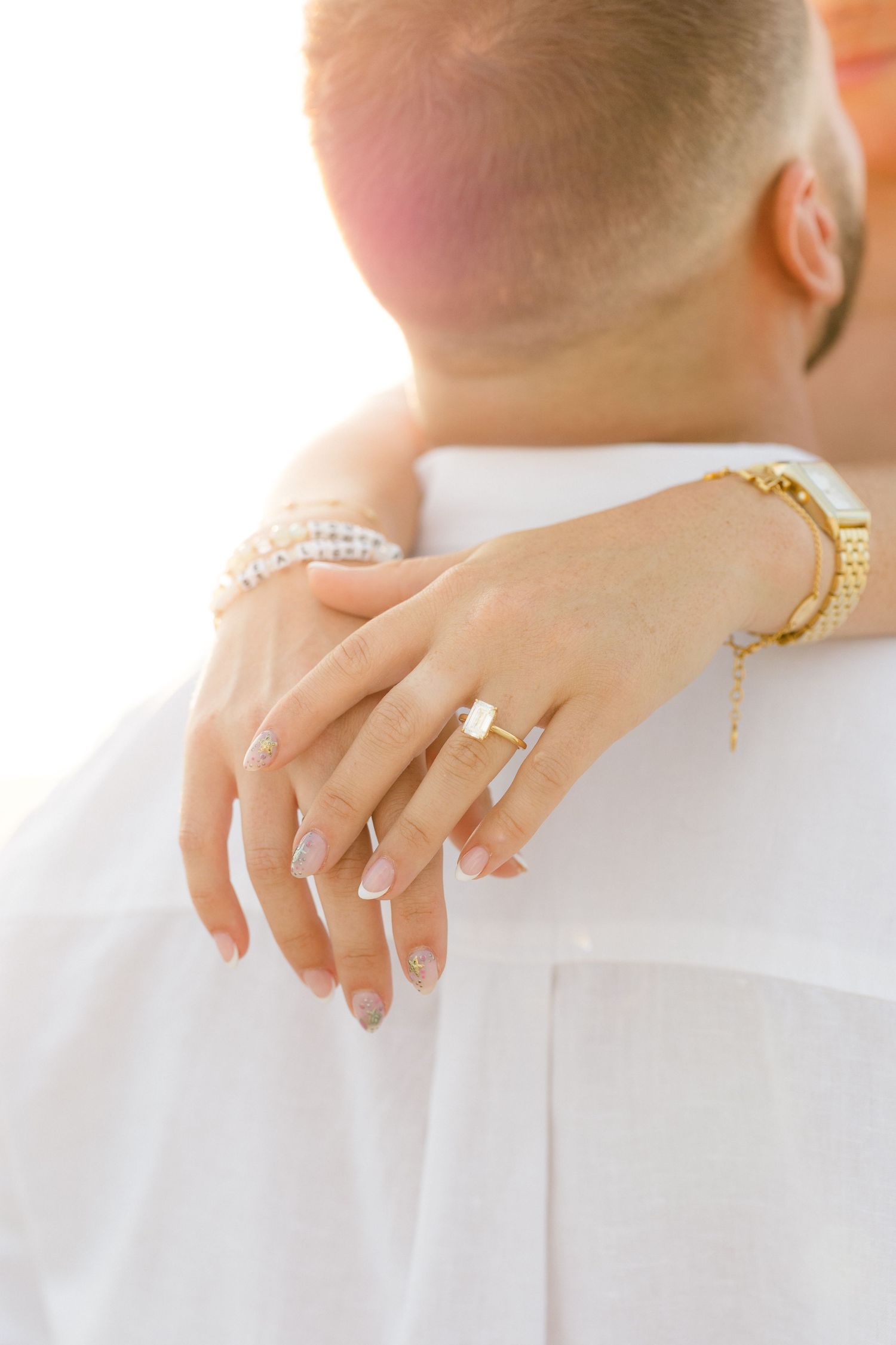 Close-up of a woman's hand with an engagement ring and gold bracelet resting on a man's white shirt shoulder.