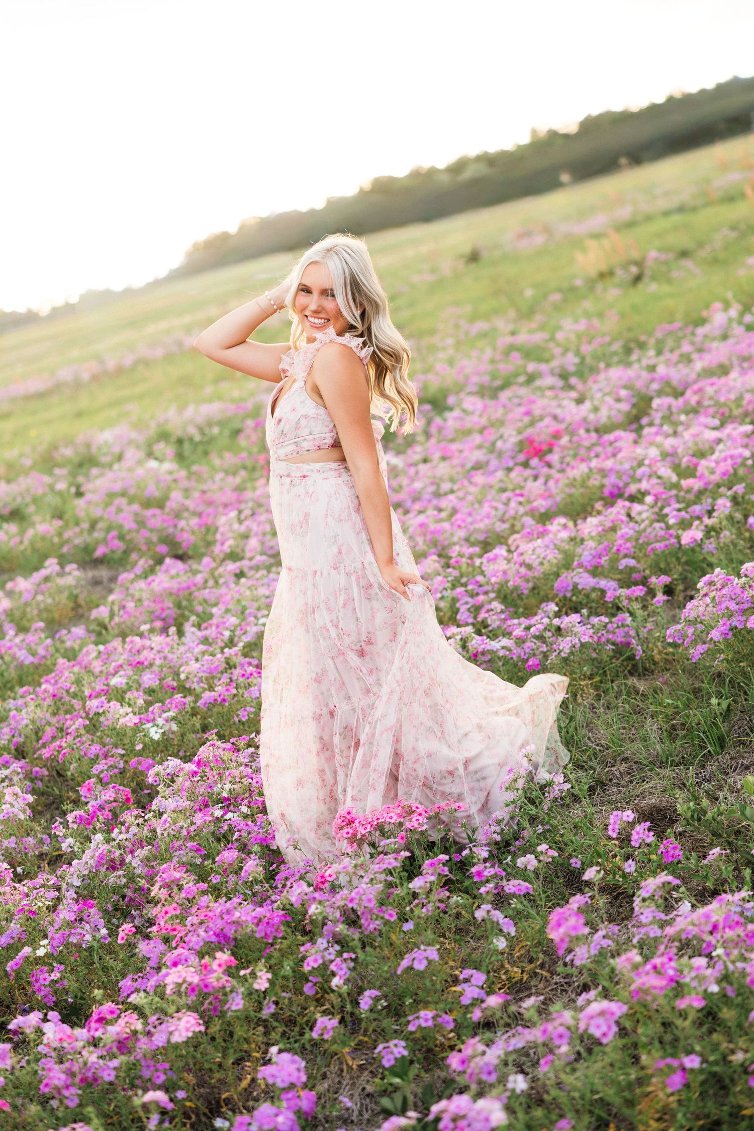 Smiling blonde woman in floral pink two-piece dress twirling in a vibrant field of purple wildflowers at golden hour.