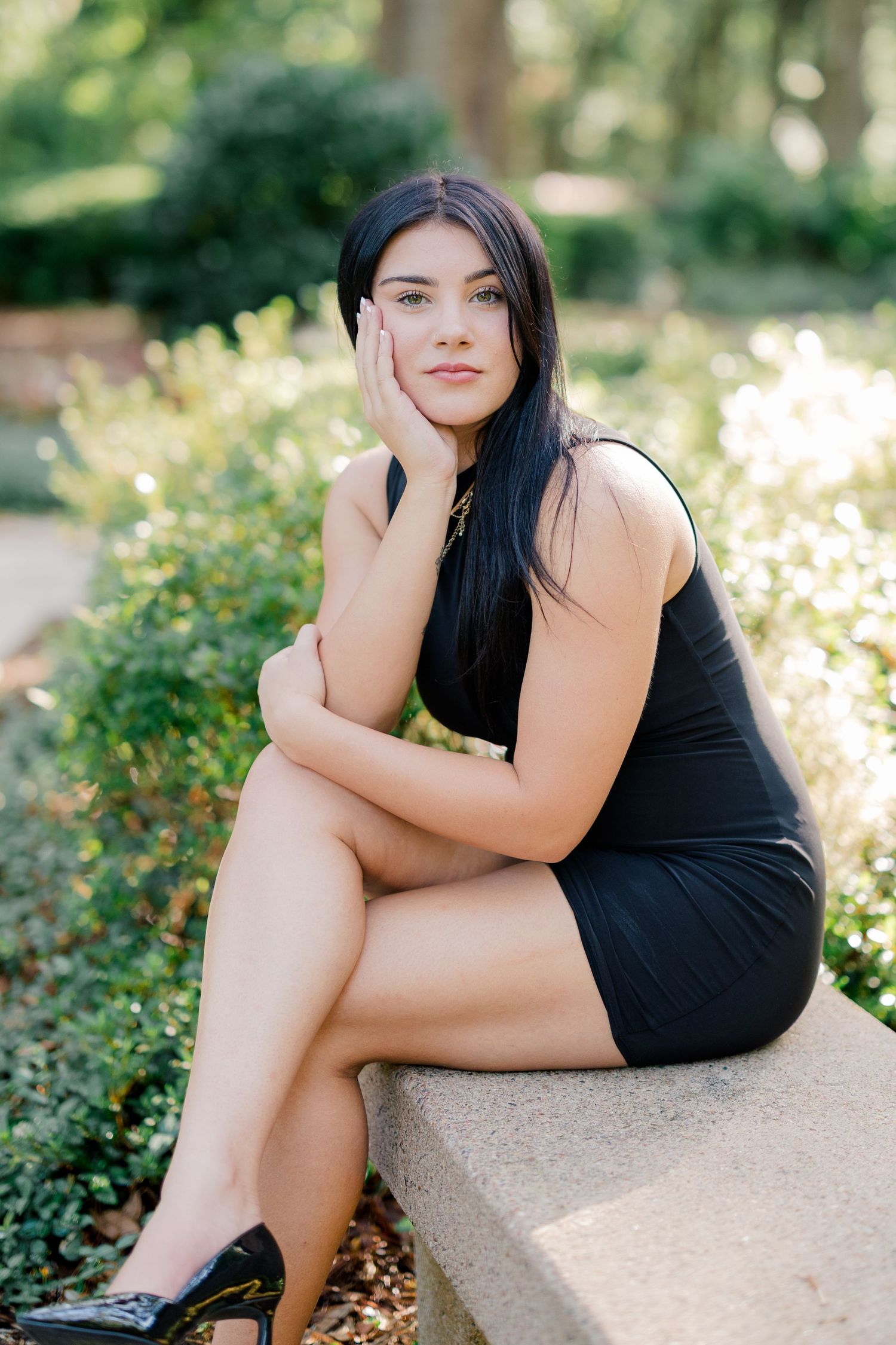 Young woman in black mini dress and heels sits on stone ledge in lush green park, posing gracefully.