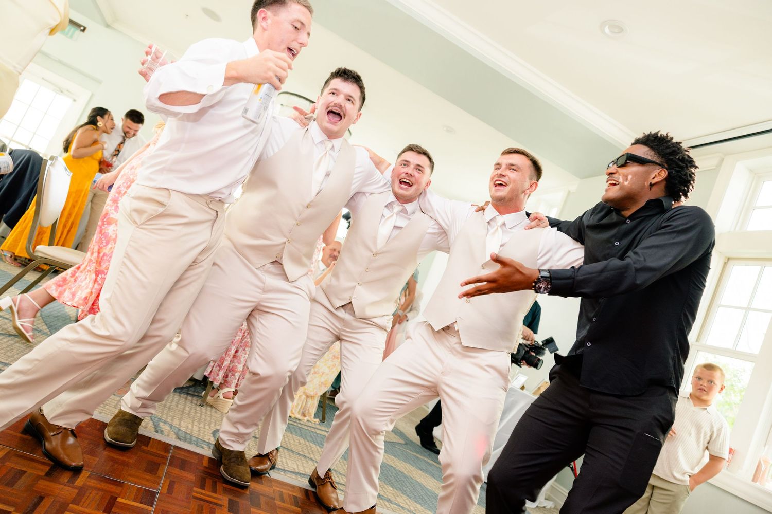 Groomsmen in white suits dancing joyfully with a DJ at a lively wedding reception celebration.
