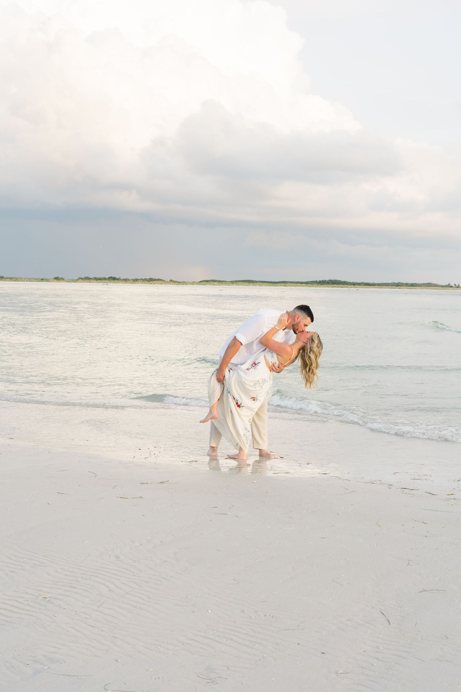 Couple sharing a romantic dip kiss on a white sand beach with ocean waves and cloudy sky backdrop.