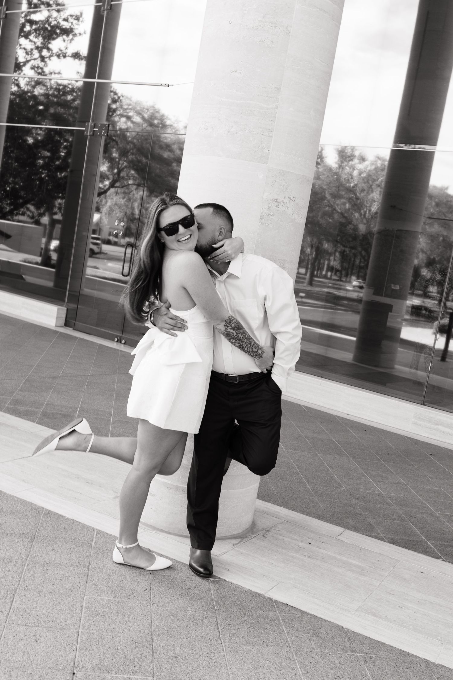 Black and white photo of a couple embracing outside a modern building, man kissing woman in white dress wearing sunglasses.