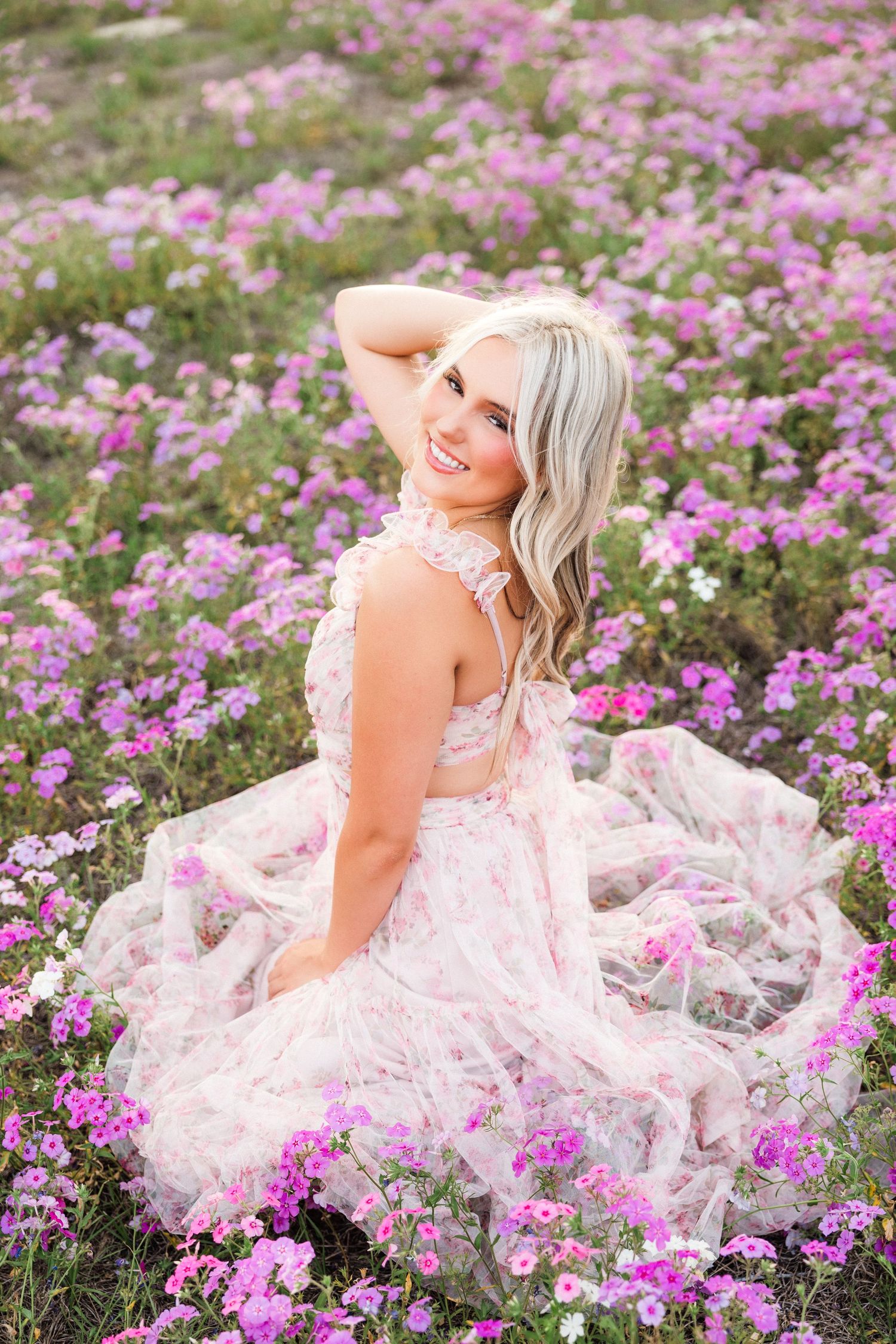 Smiling blonde woman in pink floral dress sitting in a field of purple and pink wildflowers, looking over her shoulder.