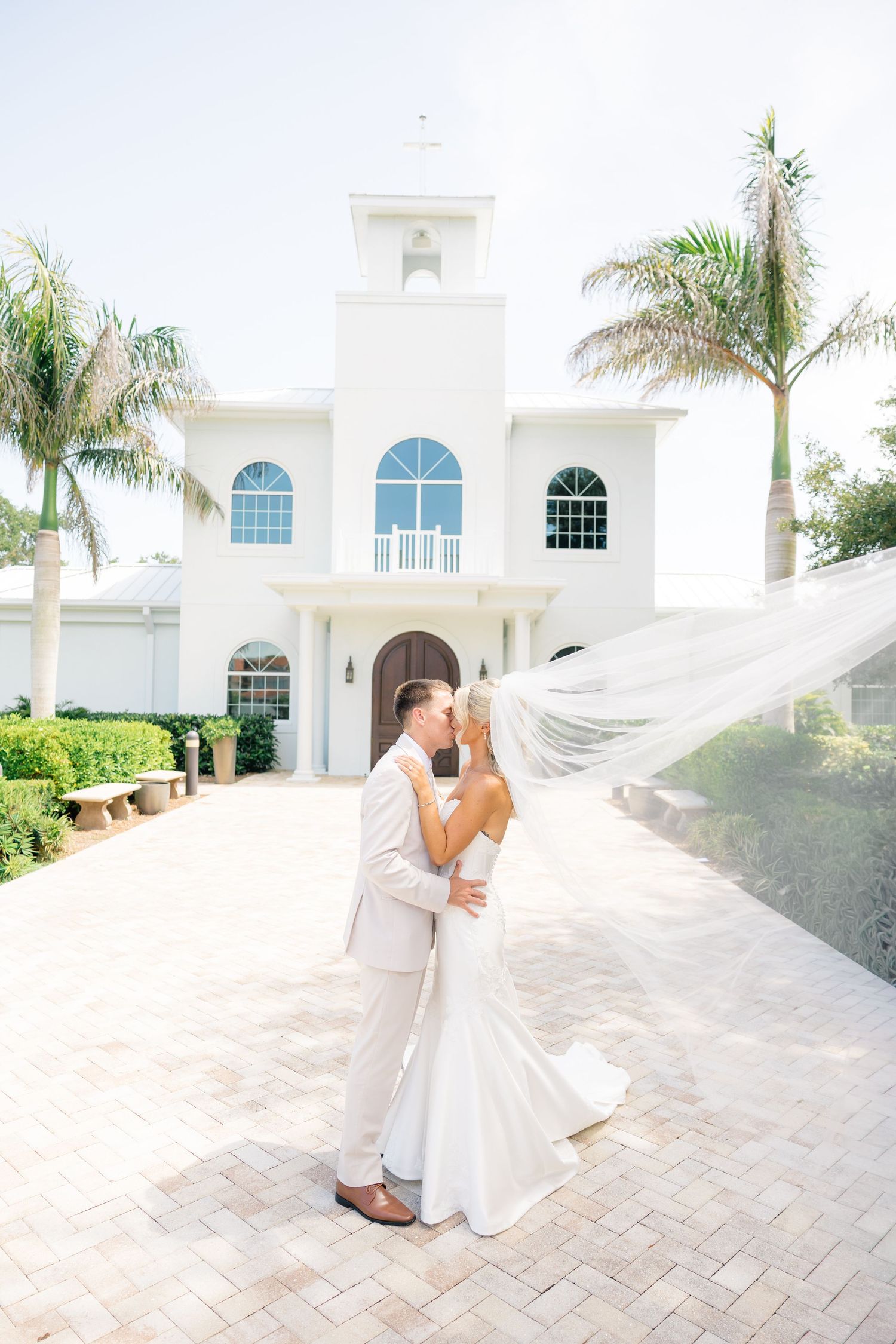Bride and groom share a kiss outside a white chapel with palm trees, bridal veil flowing in the breeze.