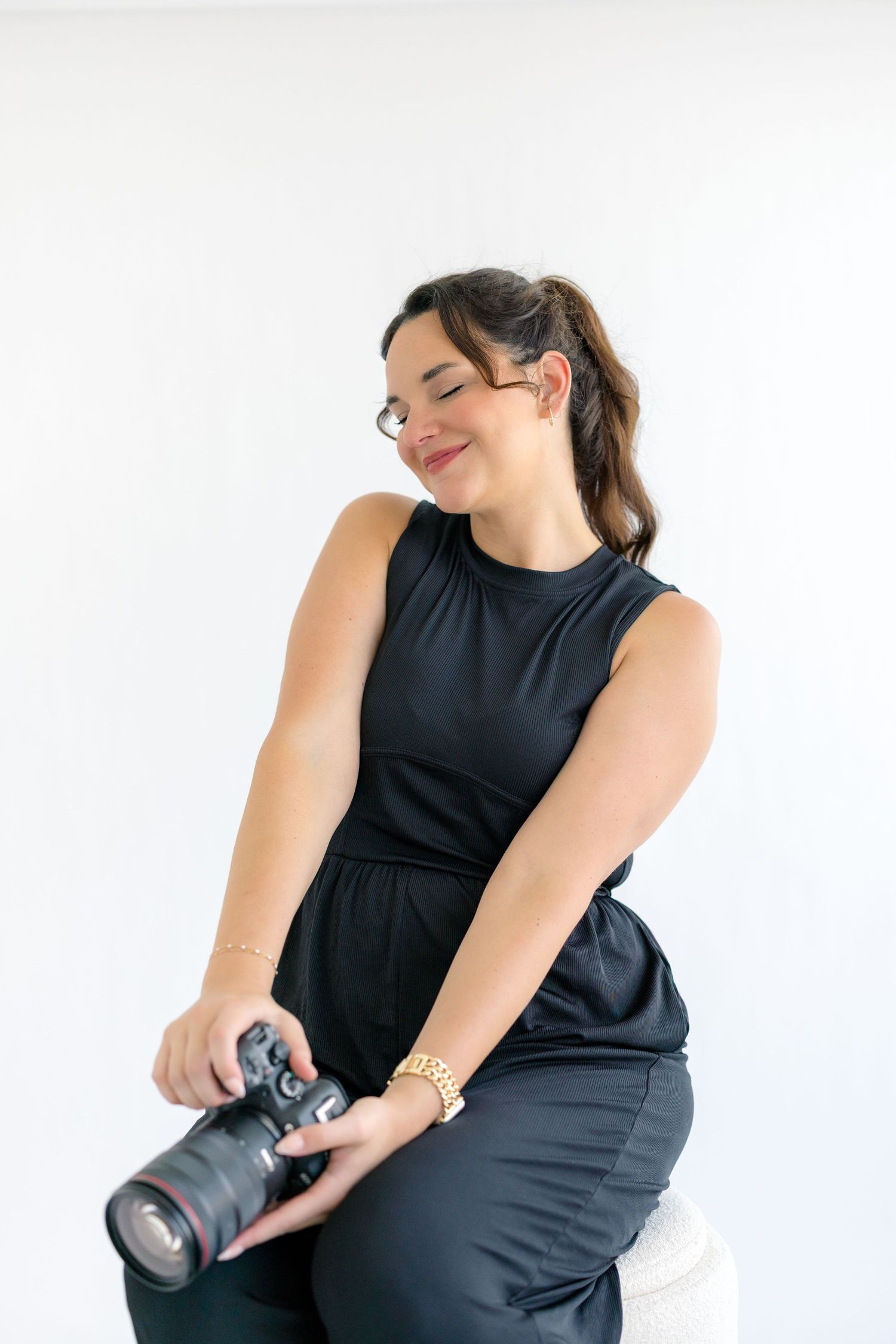 Woman in black outfit sitting and holding a DSLR camera, smiling with eyes closed against white background.