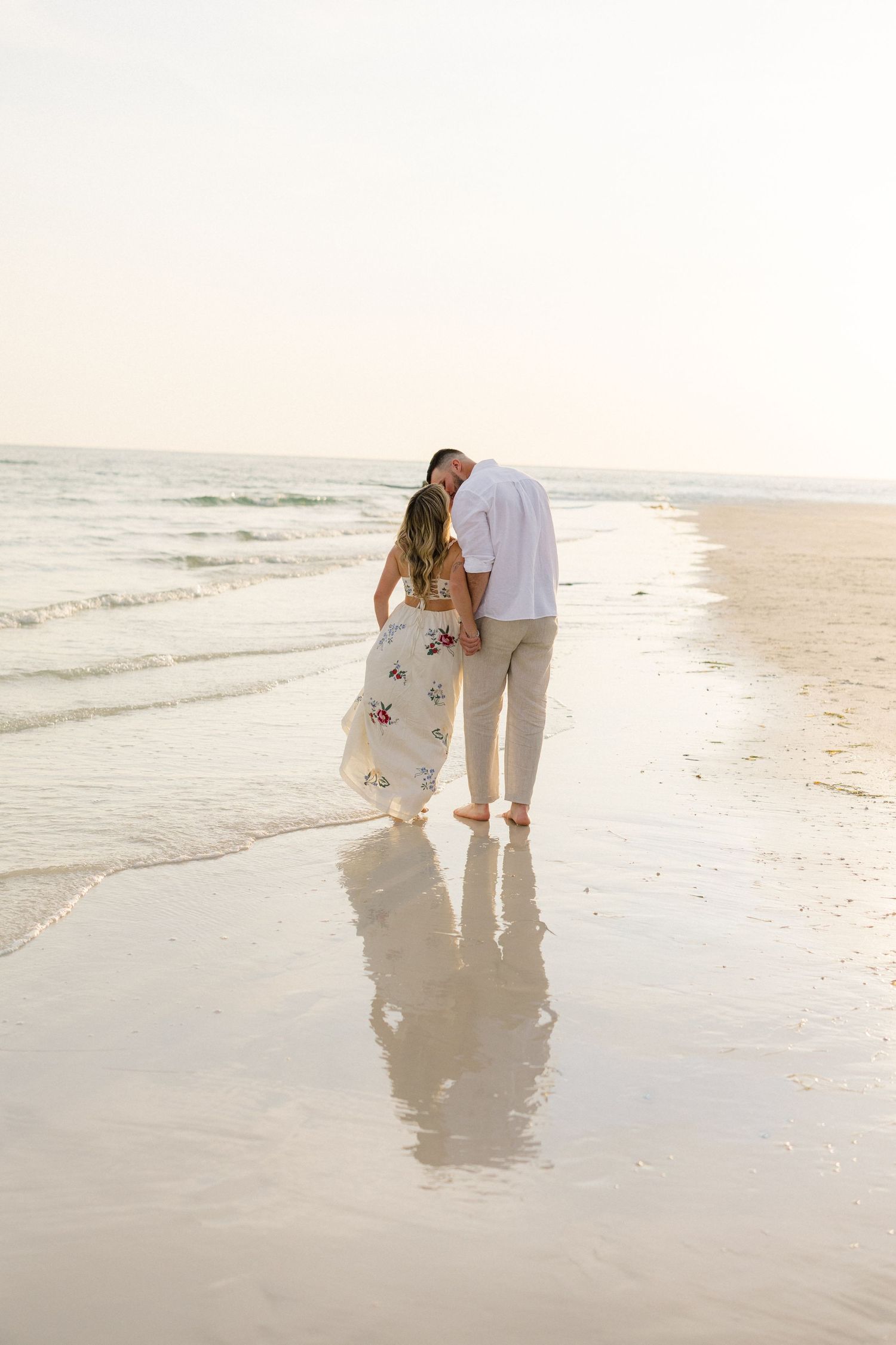 Couple walking barefoot on a sandy beach at sunset, woman in floral dress and man in white shirt, reflections in wet sand.