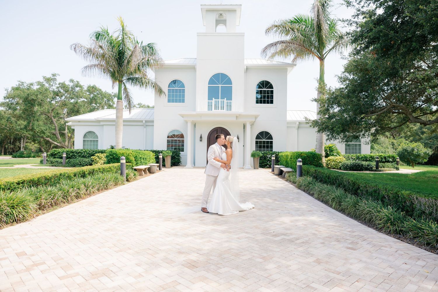 Bride and groom share a romantic kiss on a paved pathway outside a white wedding venue with palm trees.