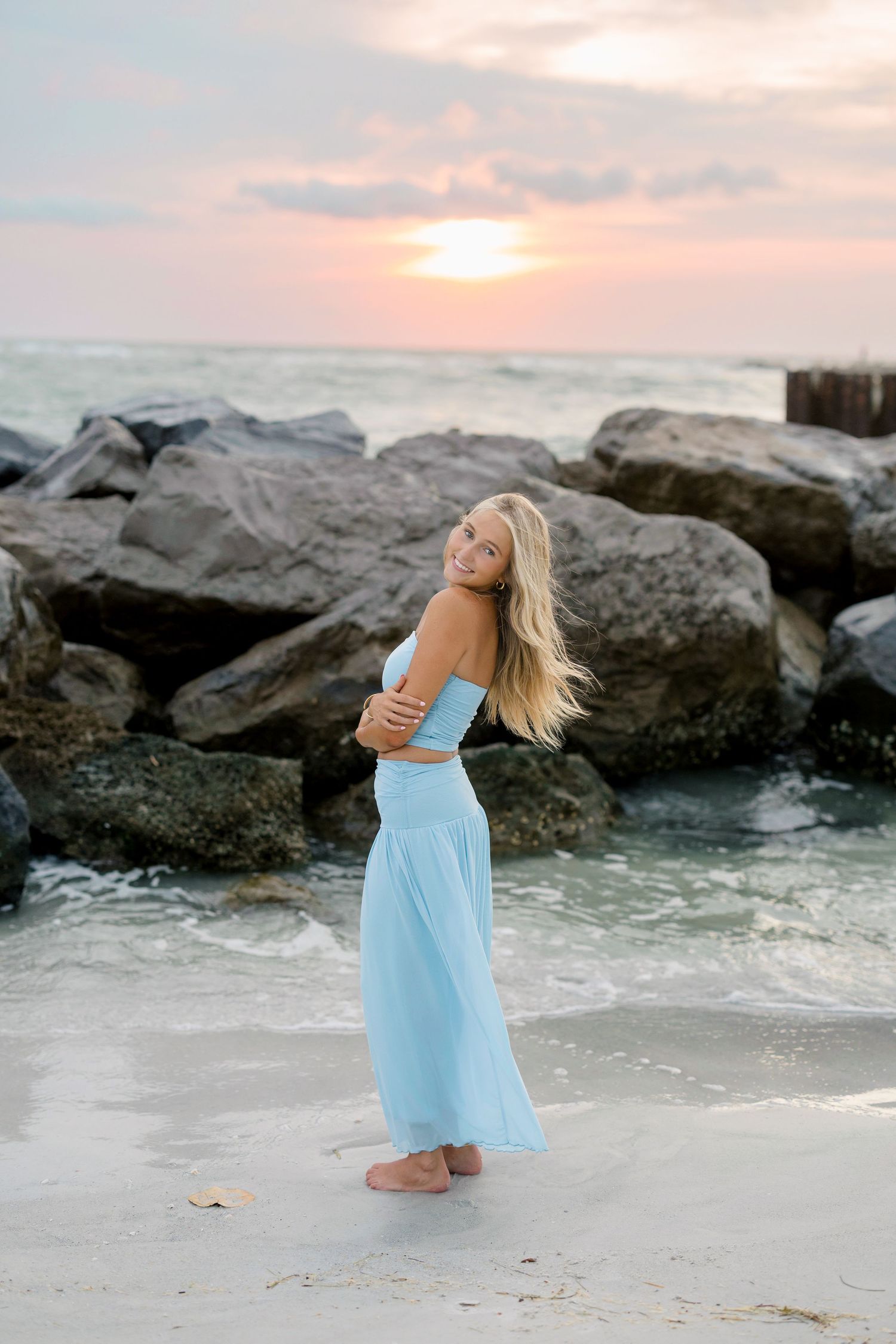 Blonde woman in light blue crop top and maxi skirt posing on a sandy beach at sunset with rocky shoreline.