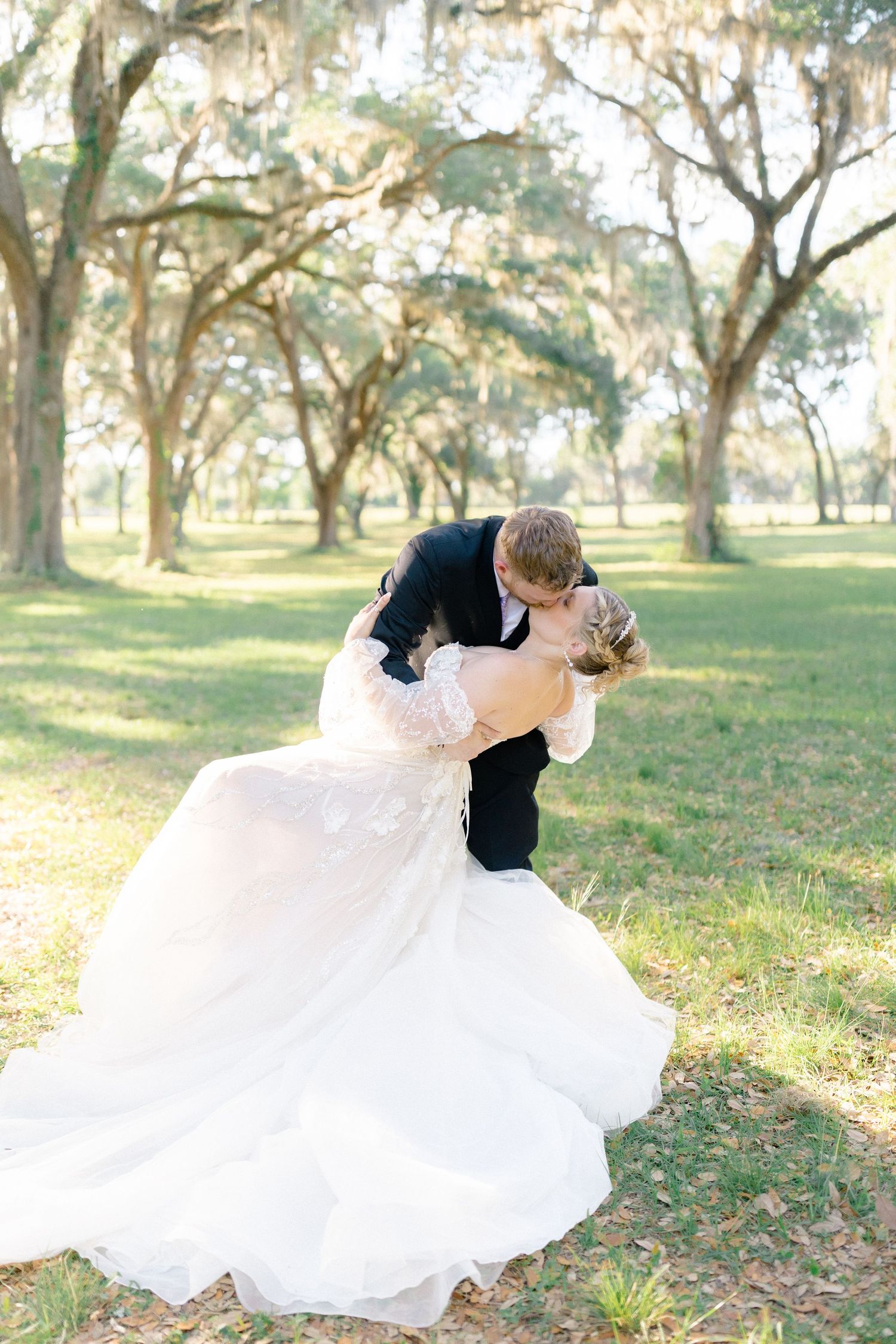 Groom dips bride for a romantic kiss under moss-draped oak trees in a sunlit outdoor wedding portrait.