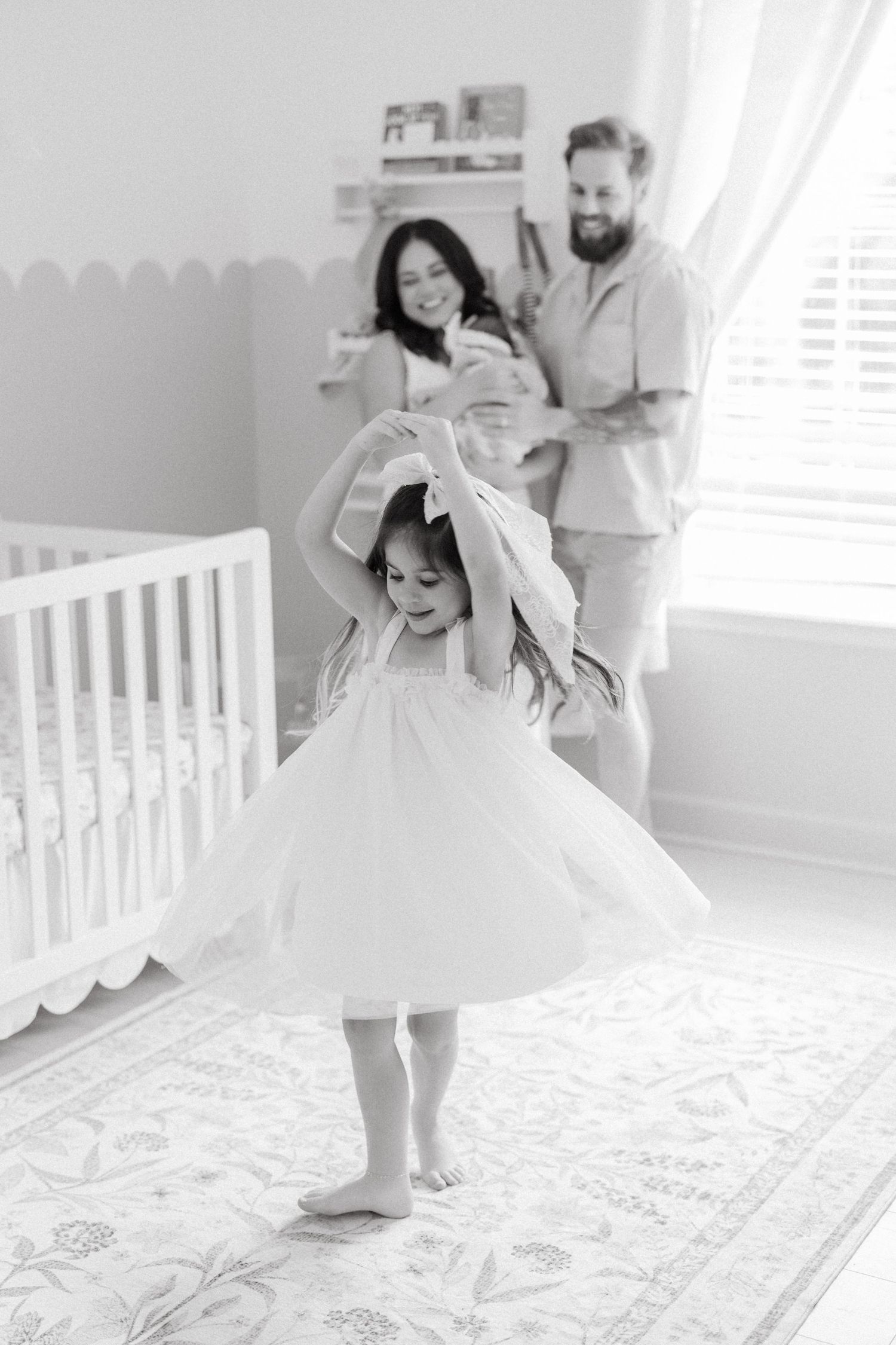 Black and white photo of a little girl in a tutu dress twirling in a nursery while her parents hold a newborn baby.