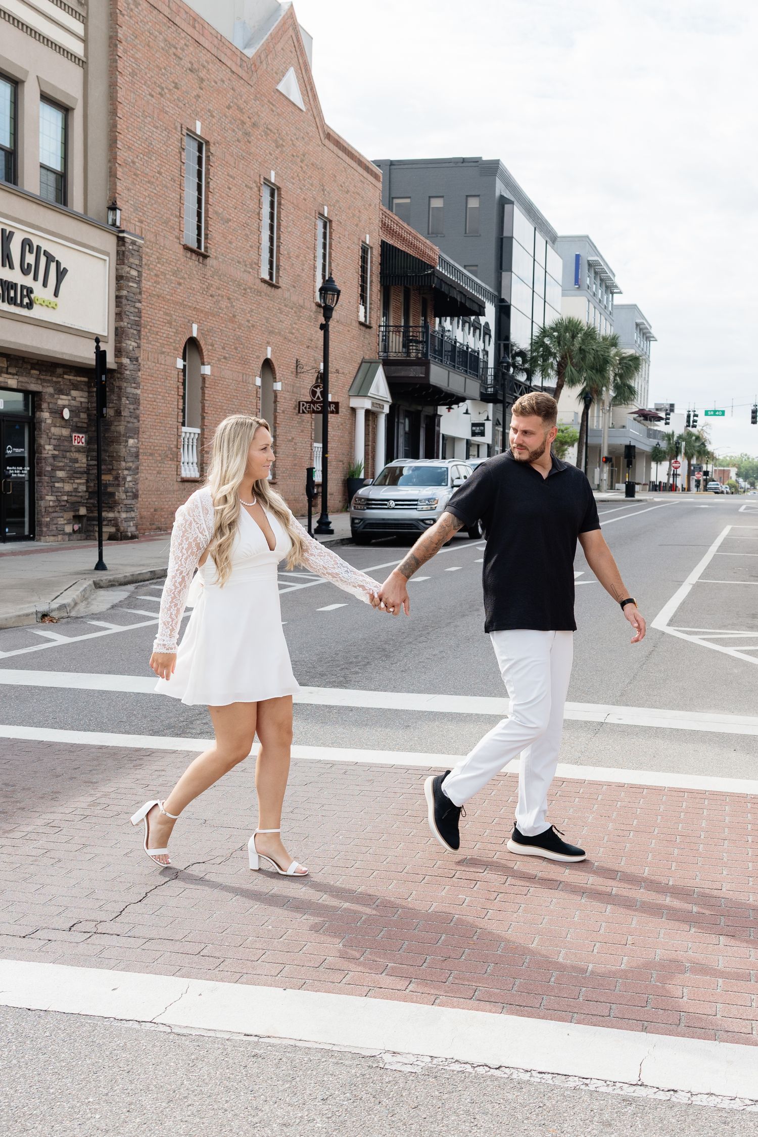 A couple holds hands while crossing a brick crosswalk in a downtown area, dressed in white and black outfits.