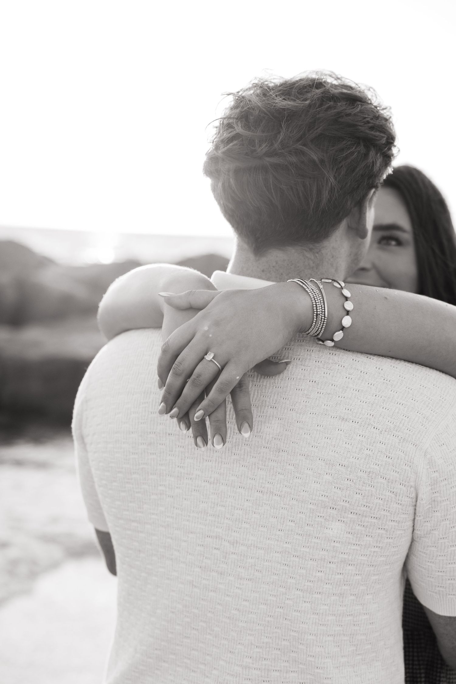Black and white photo of a couple embracing, woman's hand with engagement ring and bracelets resting on man's shoulder.