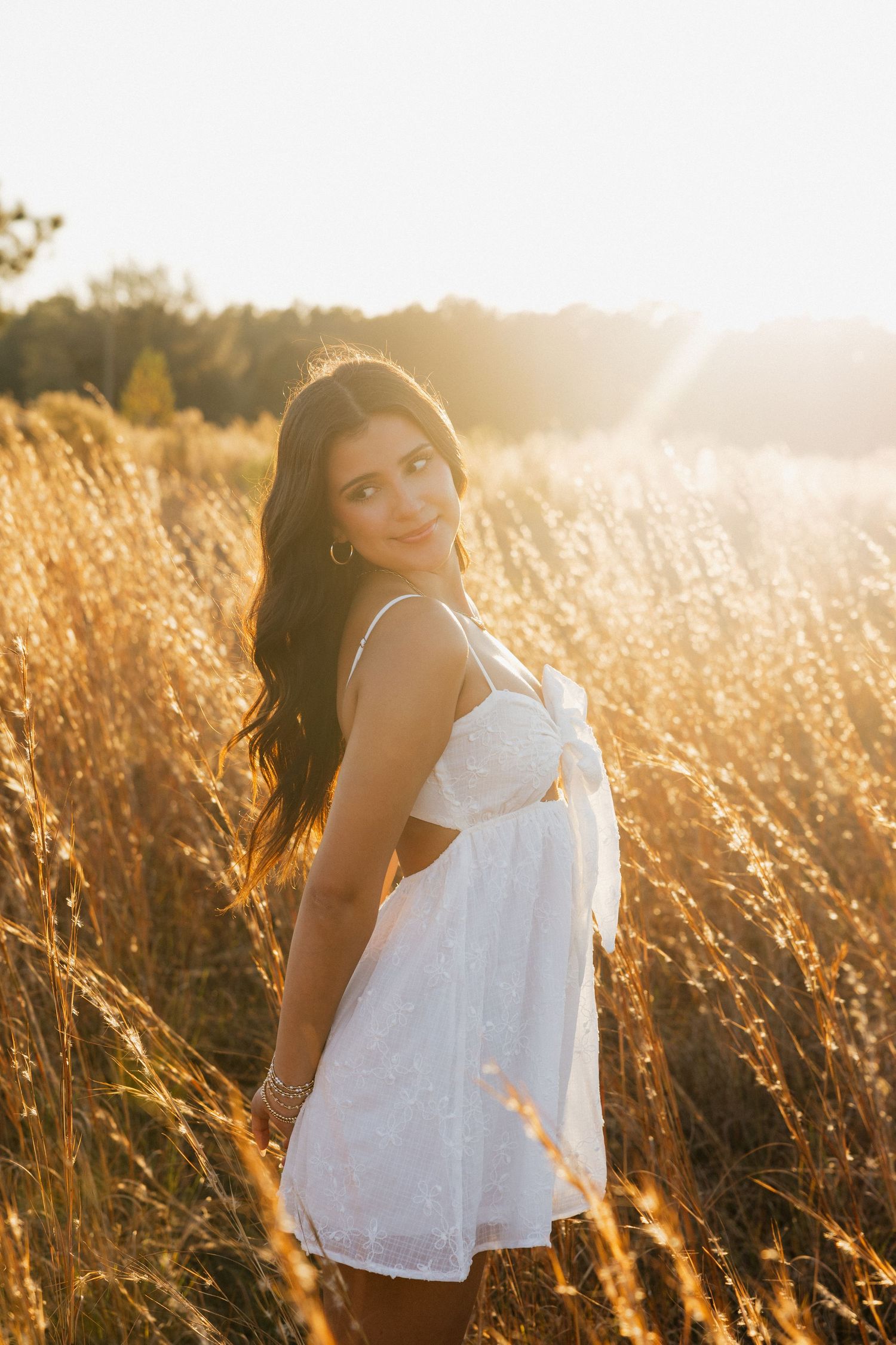 Young woman in white sundress standing in golden field at sunset with warm backlit glow.