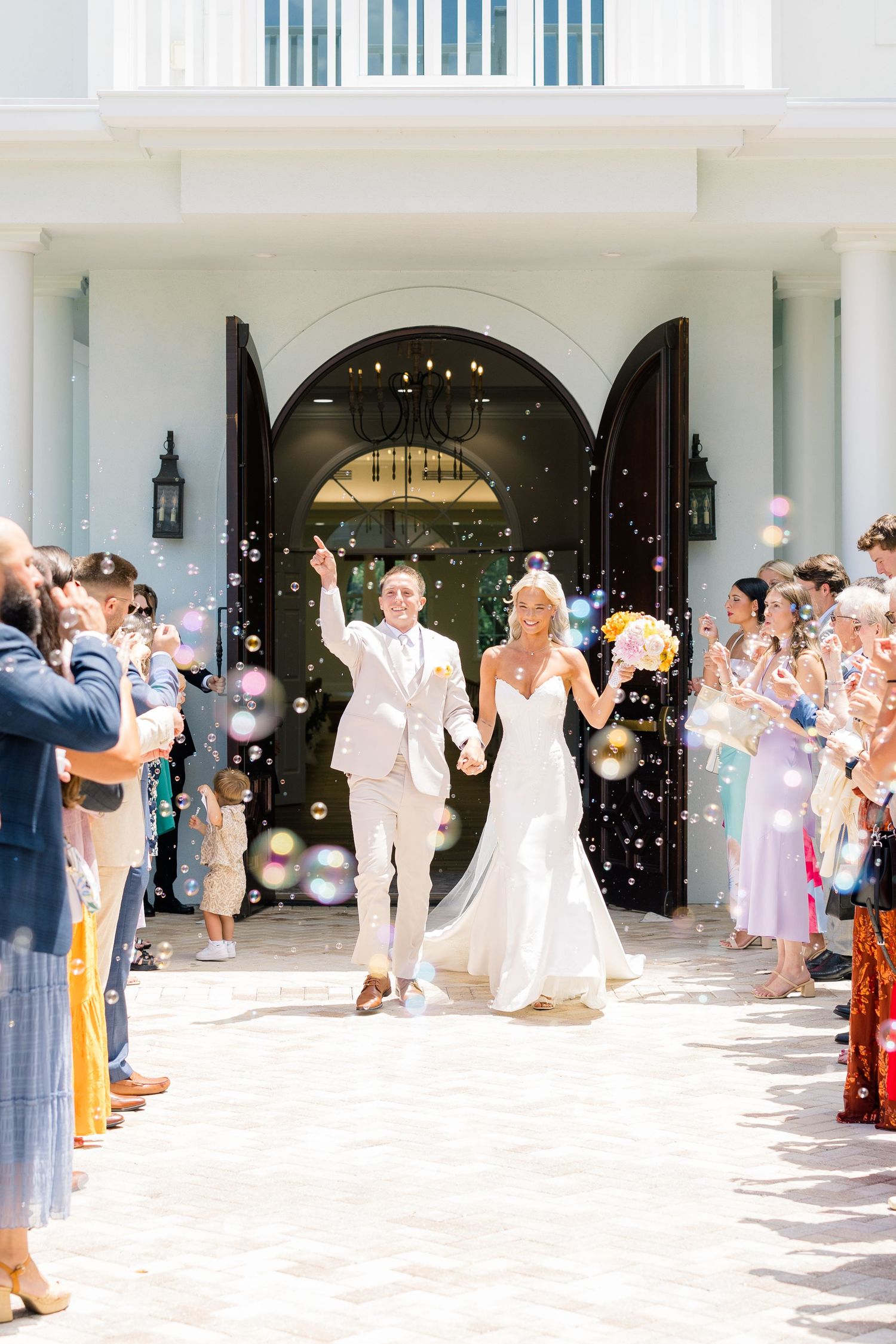 Bride and groom exit wedding venue through bubble send-off, holding hands and celebrating with guests lining the path.