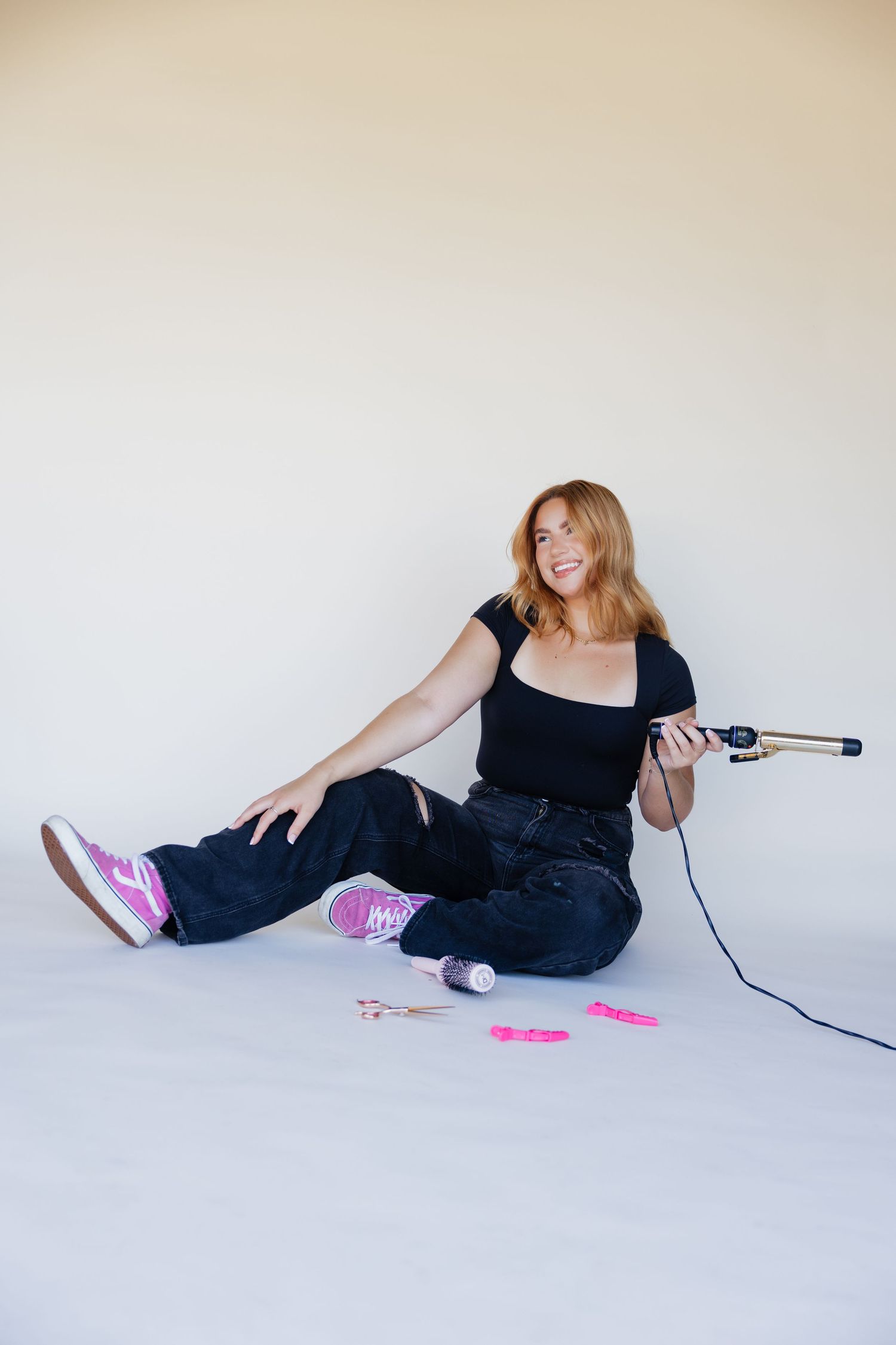 Woman sitting on floor holding curling iron, wearing black top, dark jeans, and pink sneakers, with hair clips nearby.