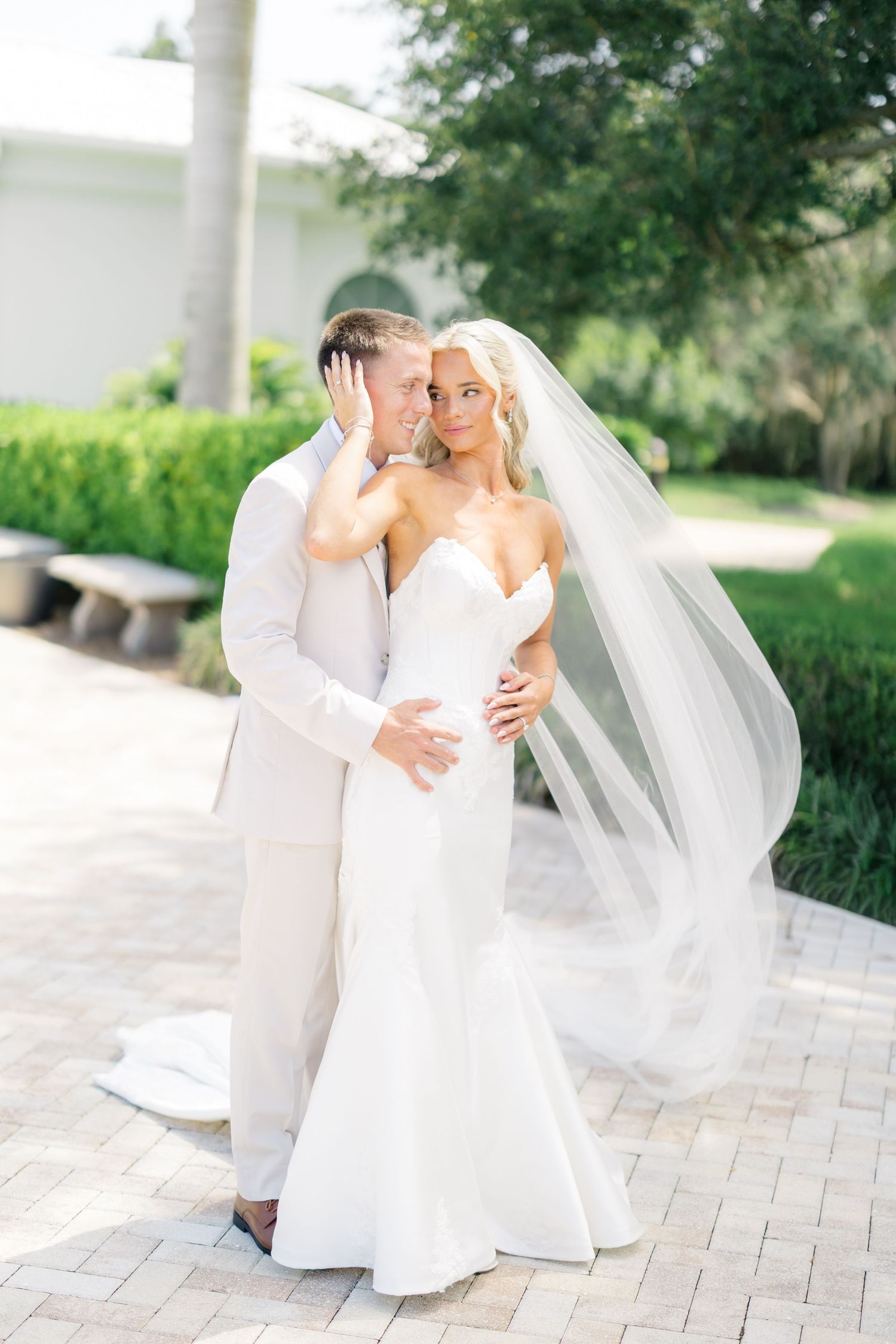 Bride in strapless mermaid gown with flowing veil embraces groom in white suit outdoors at elegant venue.
