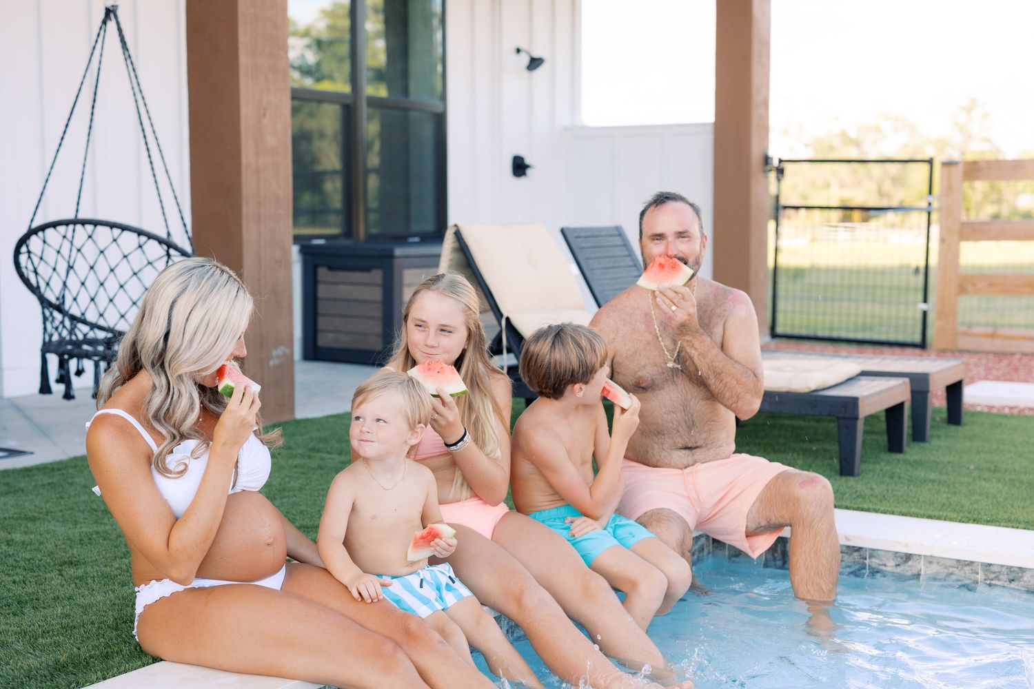 Family enjoying watermelon by a backyard pool on a sunny day, with kids and adults sitting poolside near a modern home.