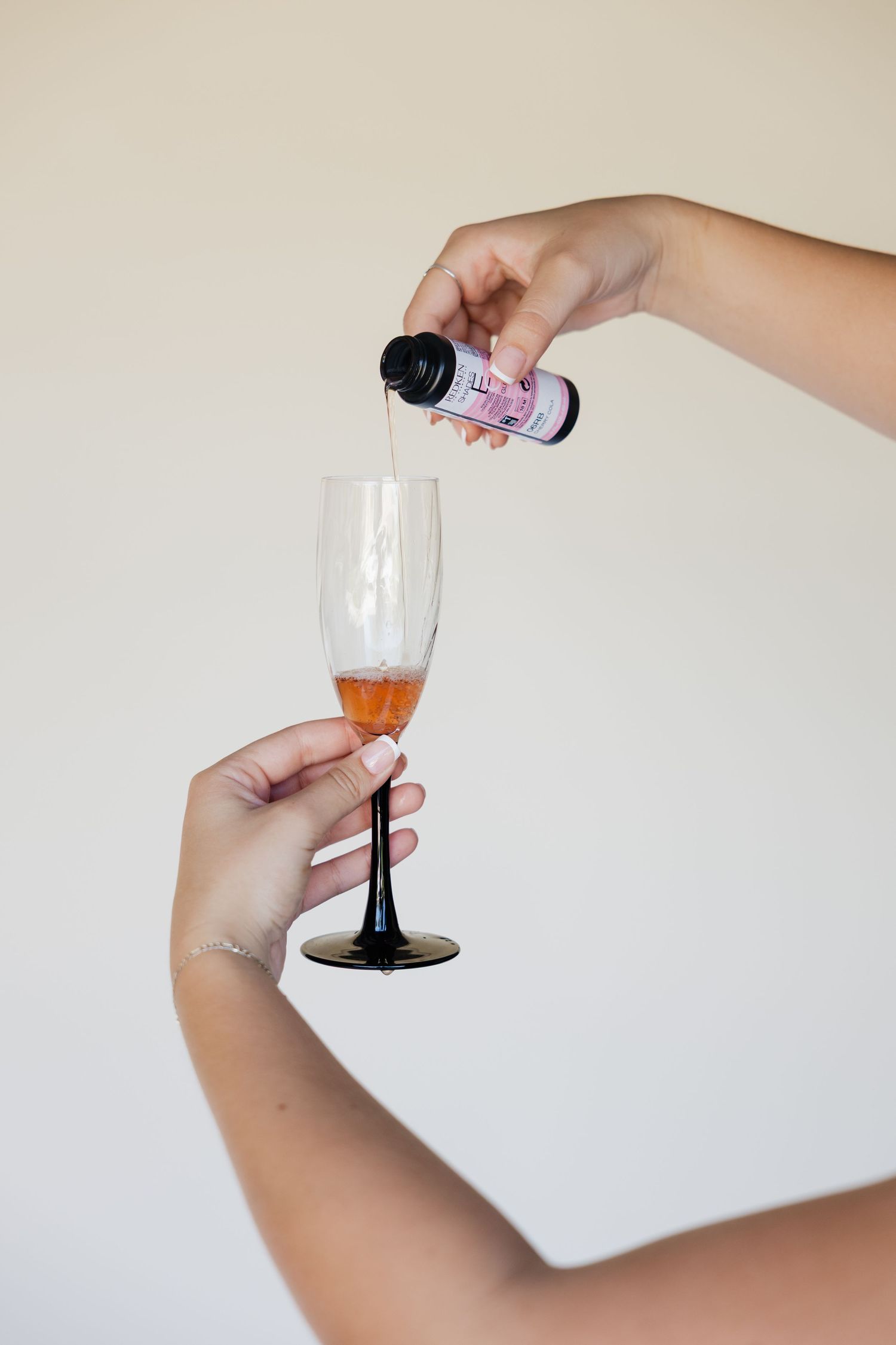 Hands pouring a pink liquid supplement into a champagne flute with a dark stem against a white background.