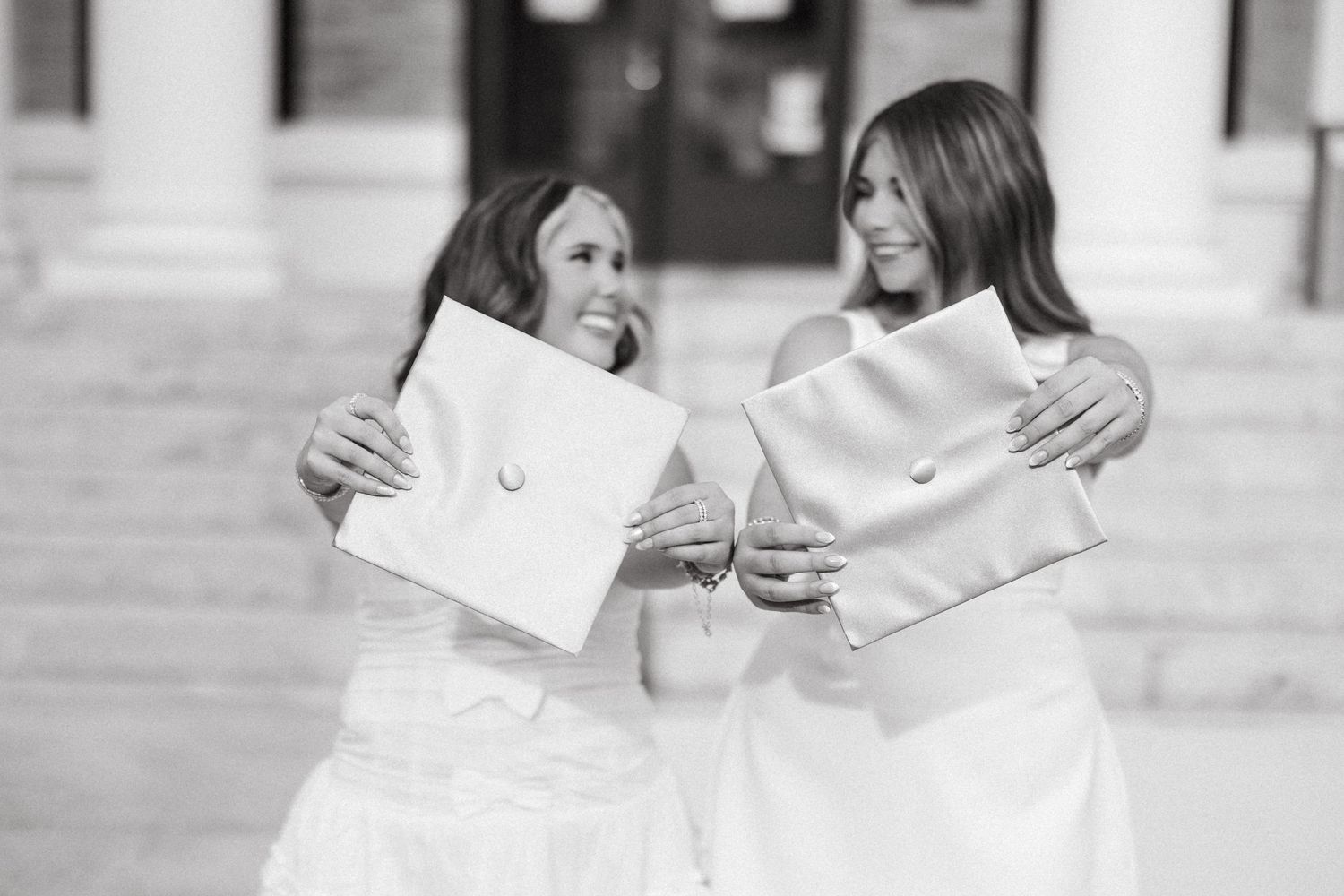 Two women in white dresses smile holding graduation caps and diplomas on steps in black and white photo.