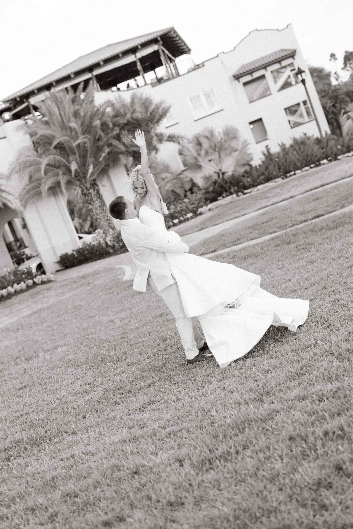 Black and white photo of groom dipping bride on a lush green lawn with palm trees.