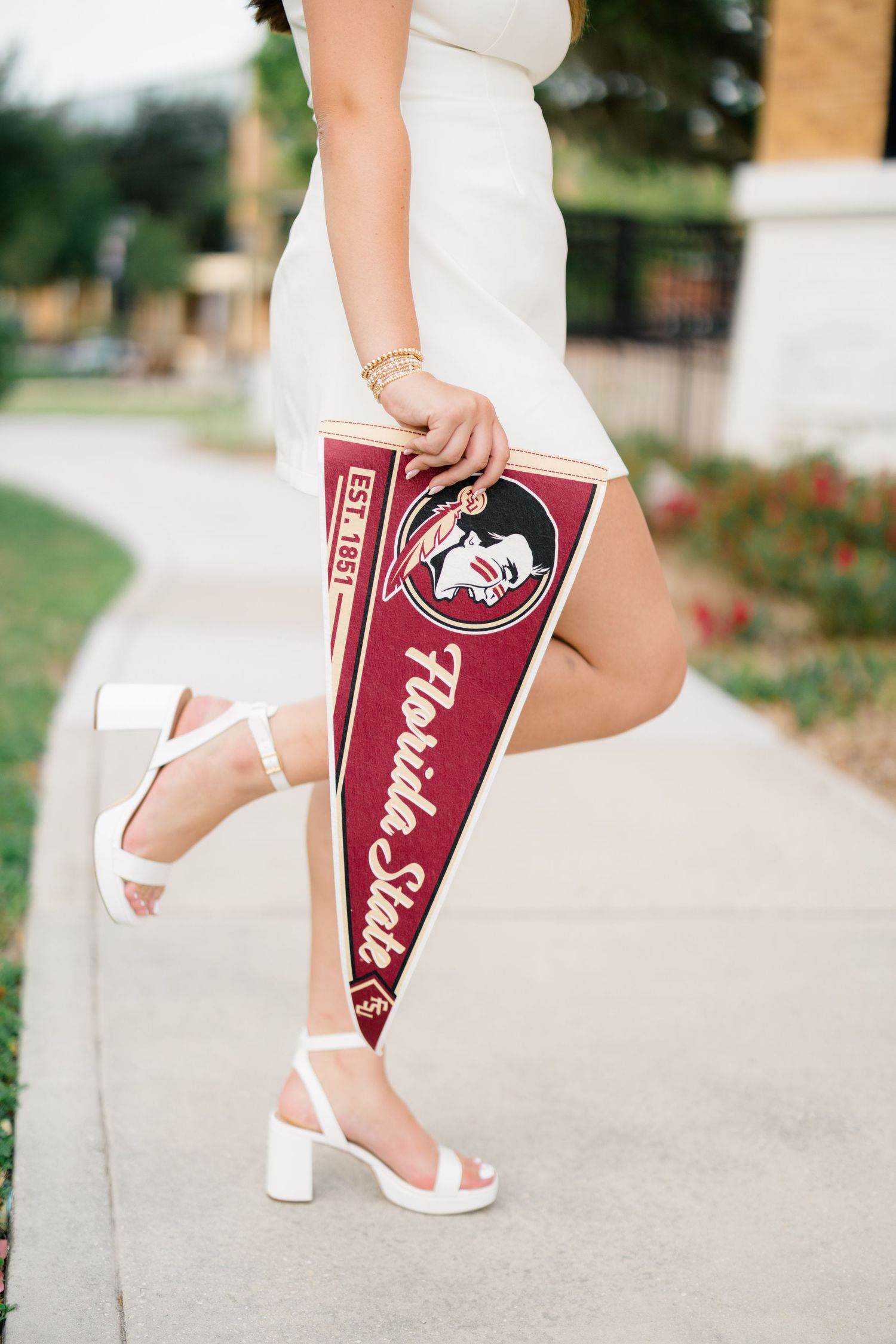 Woman in white dress holding Florida State University pennant flag, wearing white block heel sandals on a sunny sidewalk.