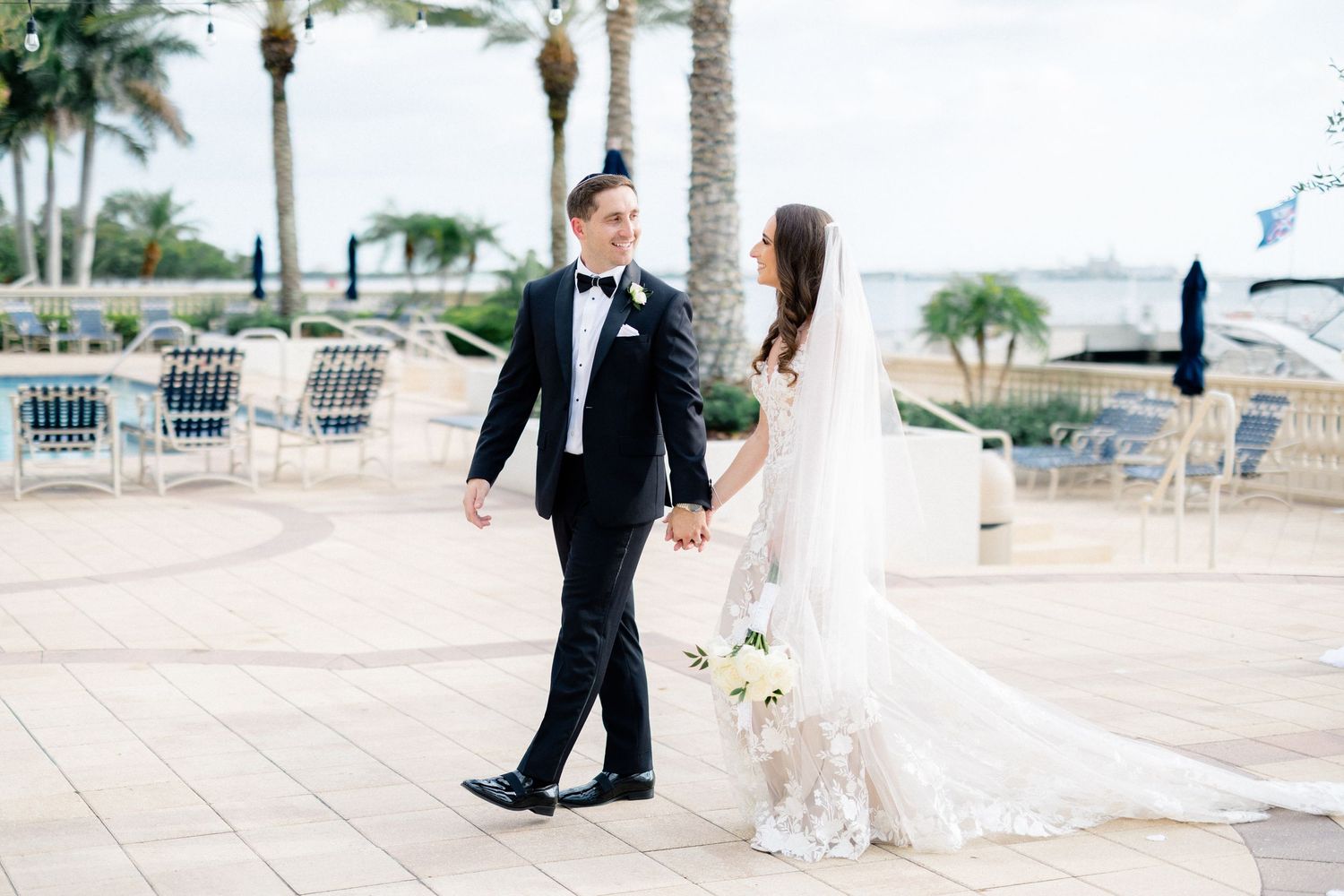 Bride and groom holding hands walking along waterfront venue with palm trees on their wedding day.