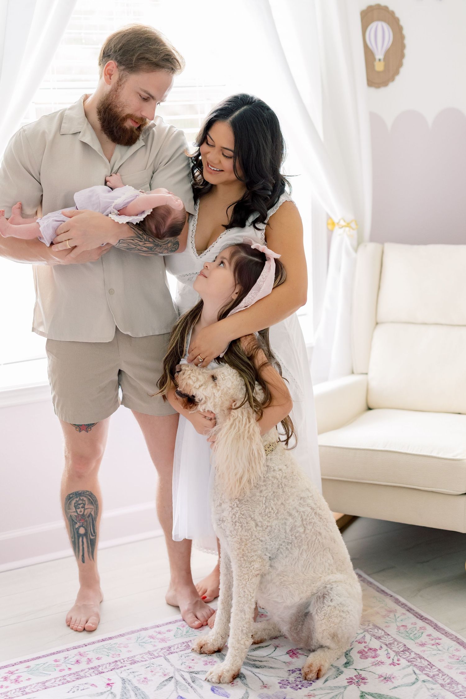 Family with newborn baby, young daughter, and large white dog posing together in a bright nursery room.