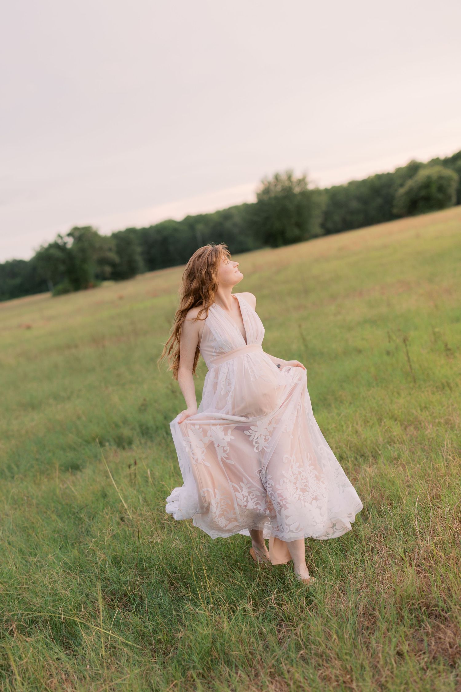 Woman in flowing lace gown twirling in green meadow at dusk.