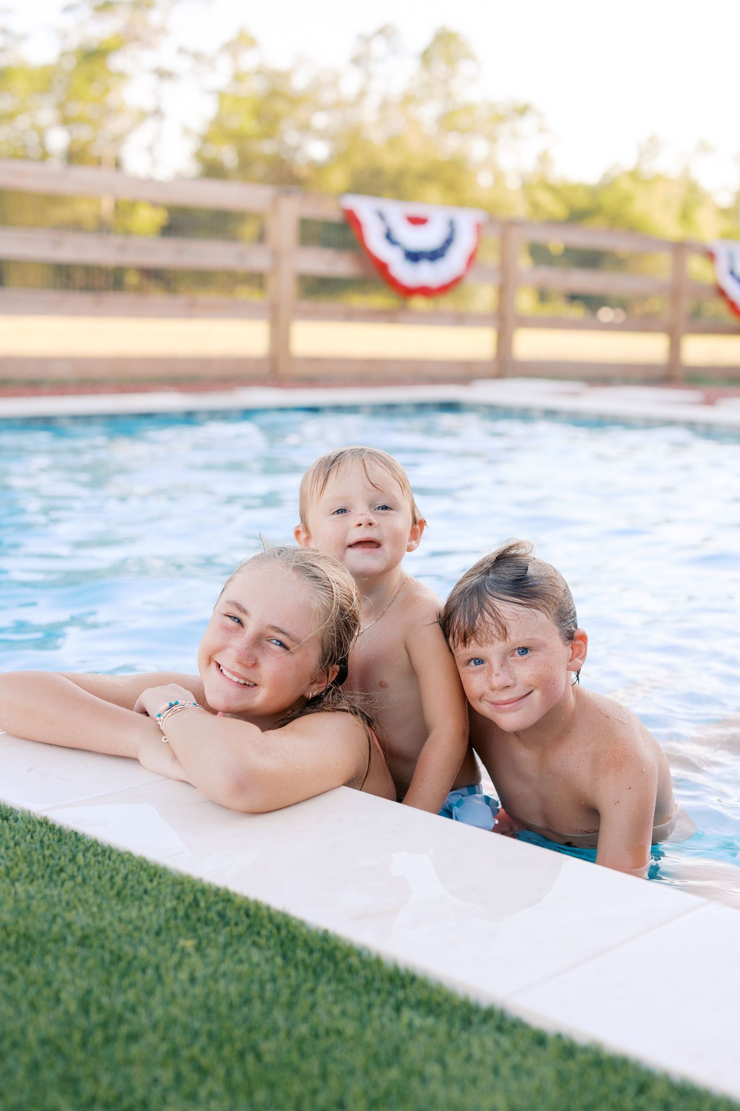 Three children smiling and posing at the edge of a backyard pool decorated with patriotic American bunting.