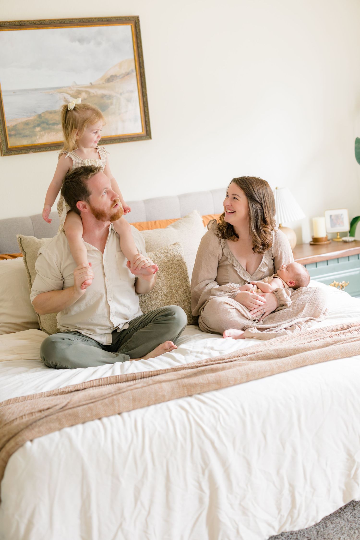 A happy family sits on a bed, dad holds toddler on shoulders while mom cradles newborn baby in a bright bedroom.