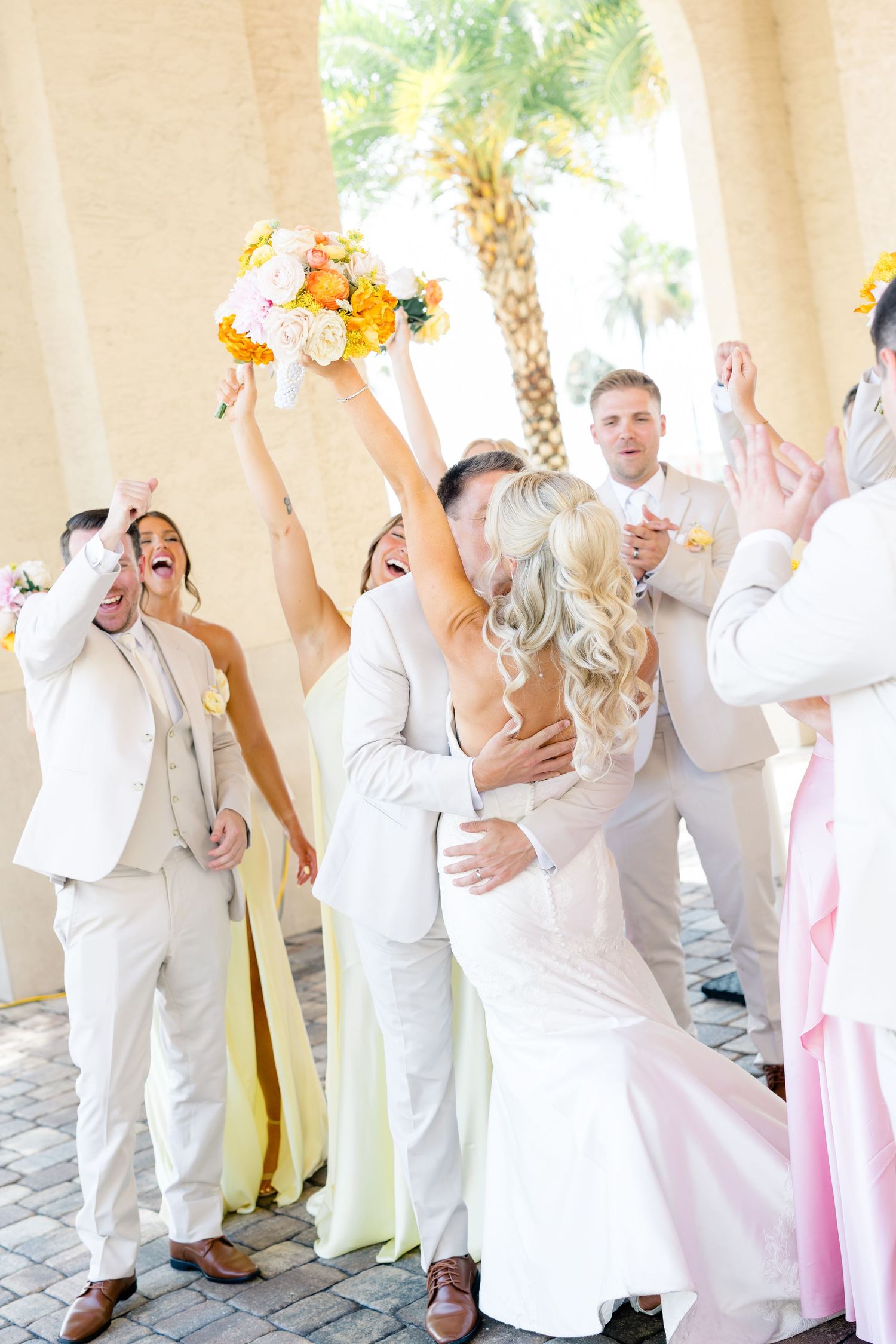Bride and groom share a first kiss surrounded by cheering wedding party in white and yellow attire outdoors.
