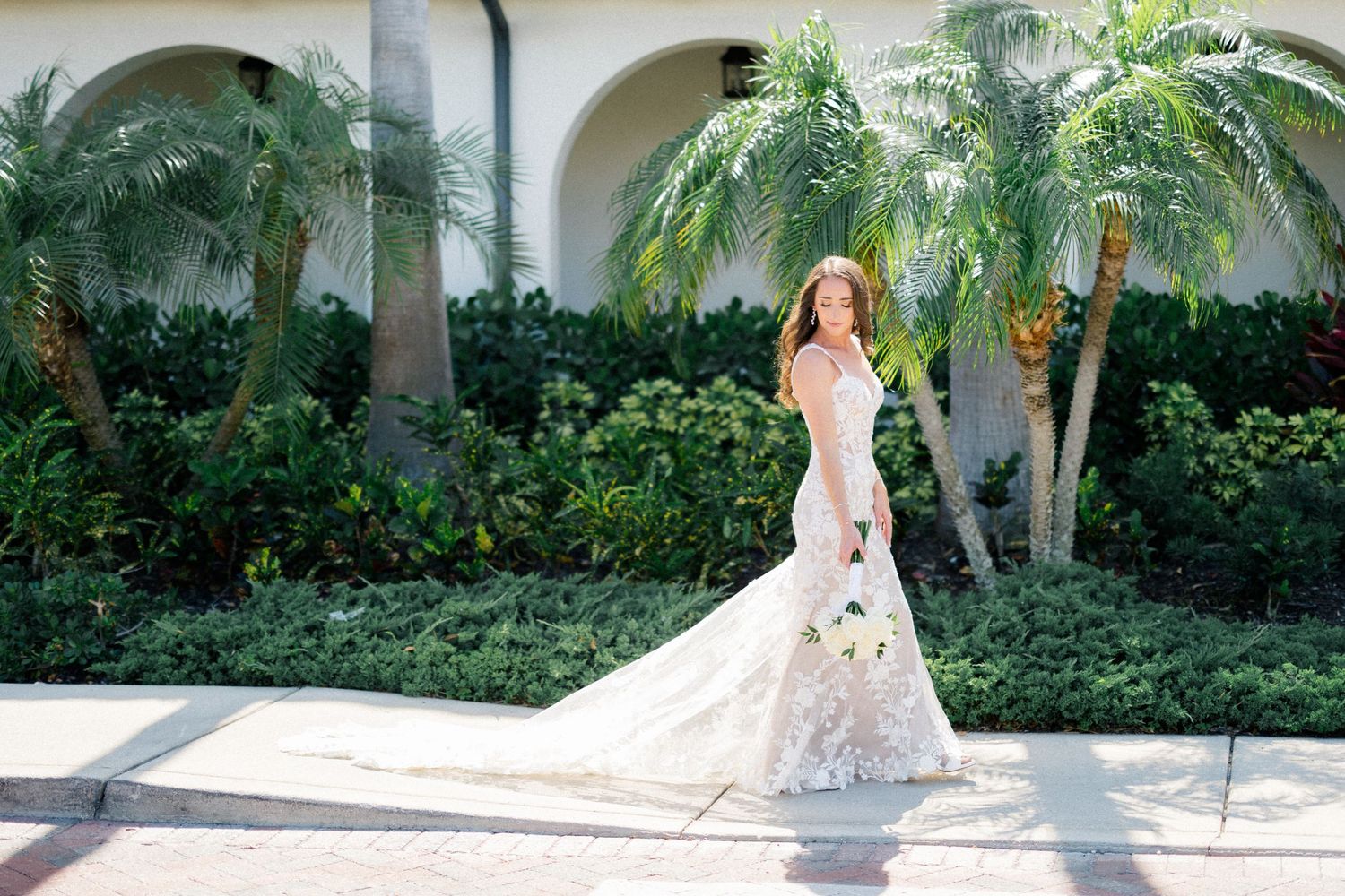 Bride in elegant lace mermaid wedding gown holding bouquet, posing outdoors near palm trees and lush greenery.