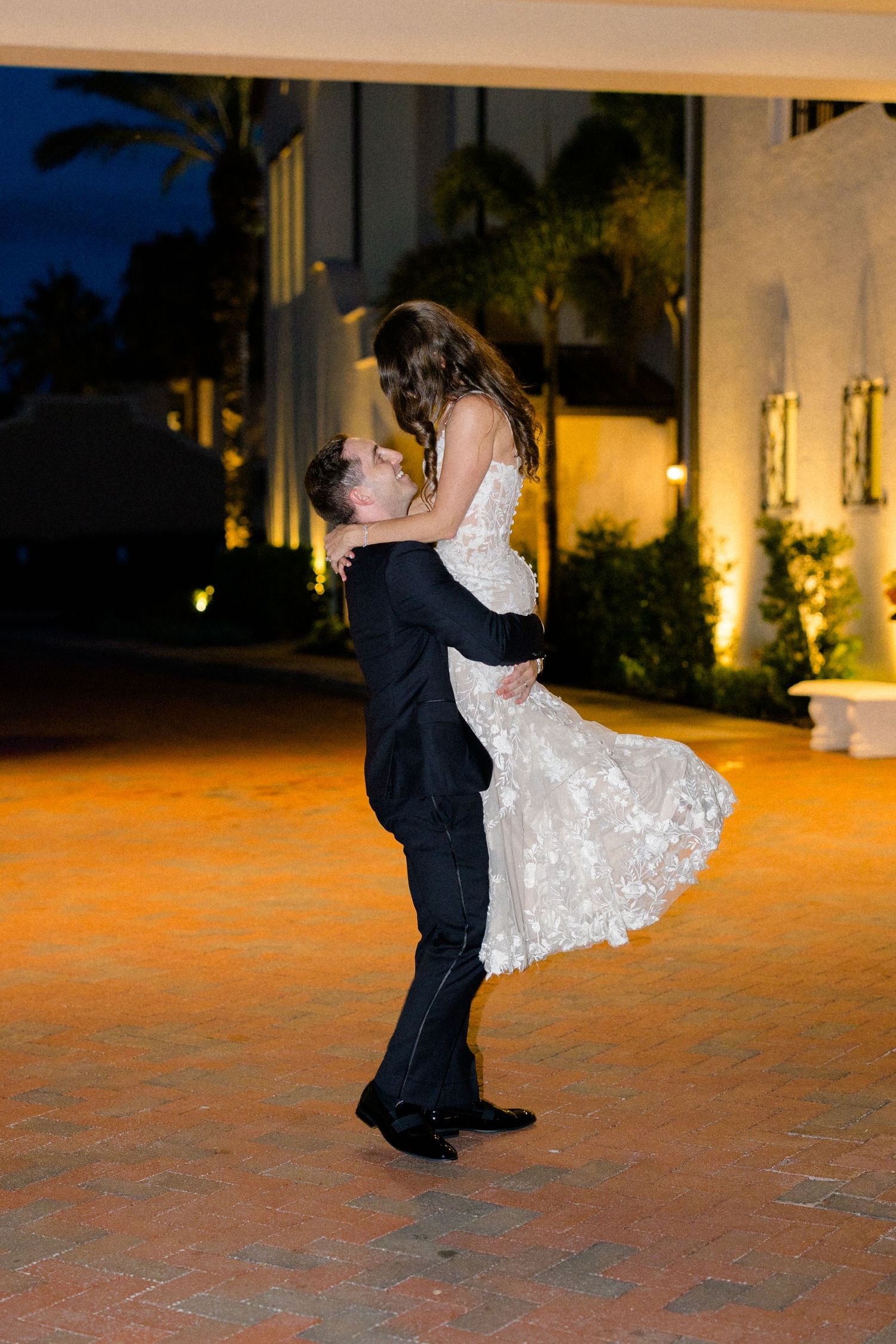 Groom lifting bride in white lace dress outside illuminated venue during romantic nighttime wedding celebration.