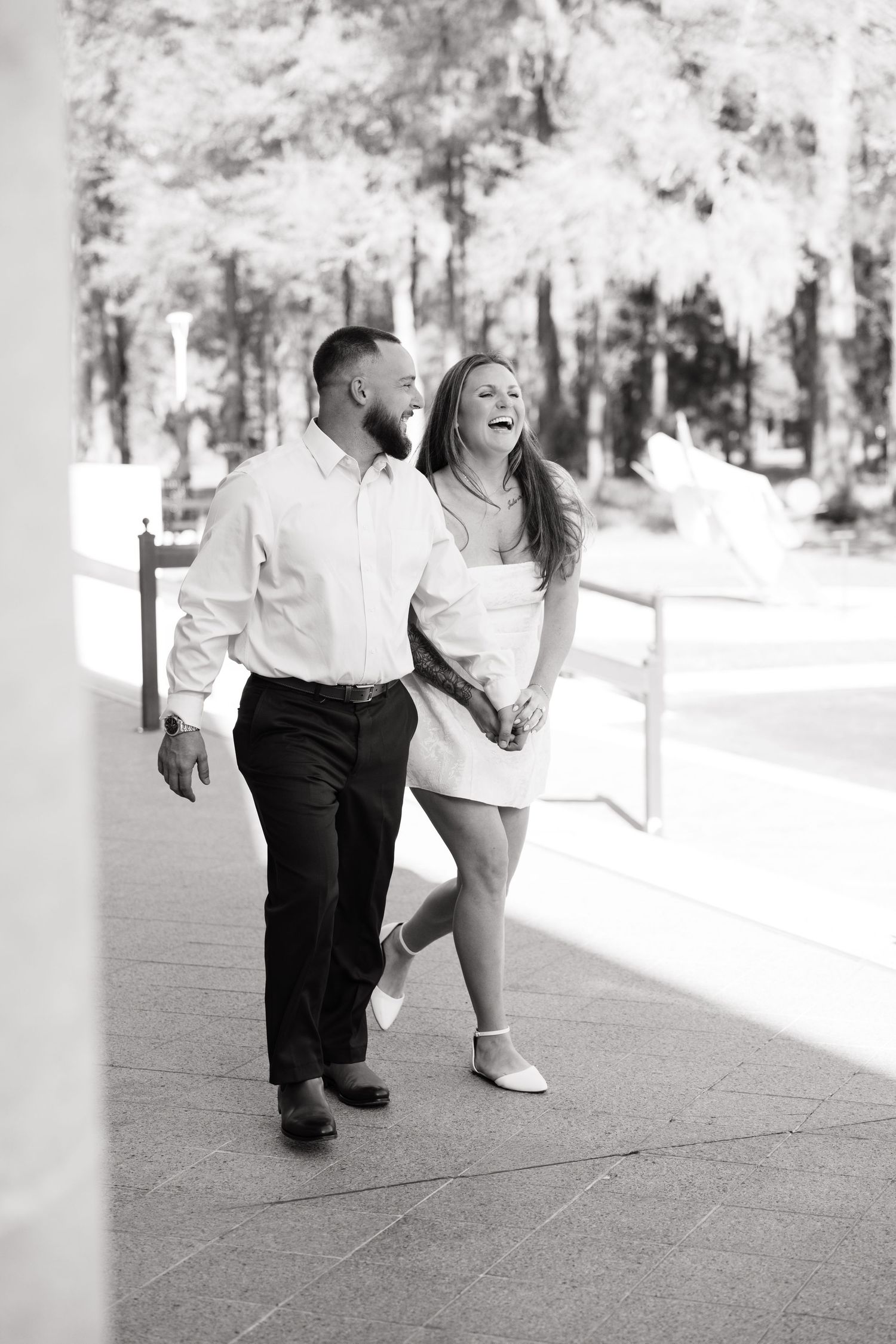 Black and white photo of a laughing couple walking hand in hand outdoors near trees and a waterfront.
