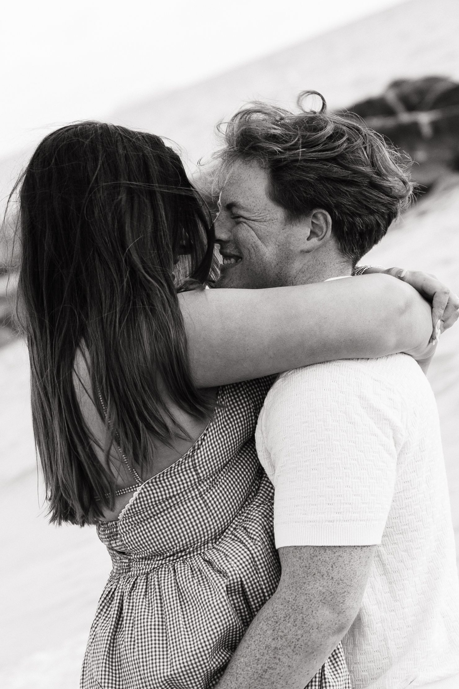 Black and white photo of a happy couple embracing and smiling on a windy beach, woman in checkered dress.