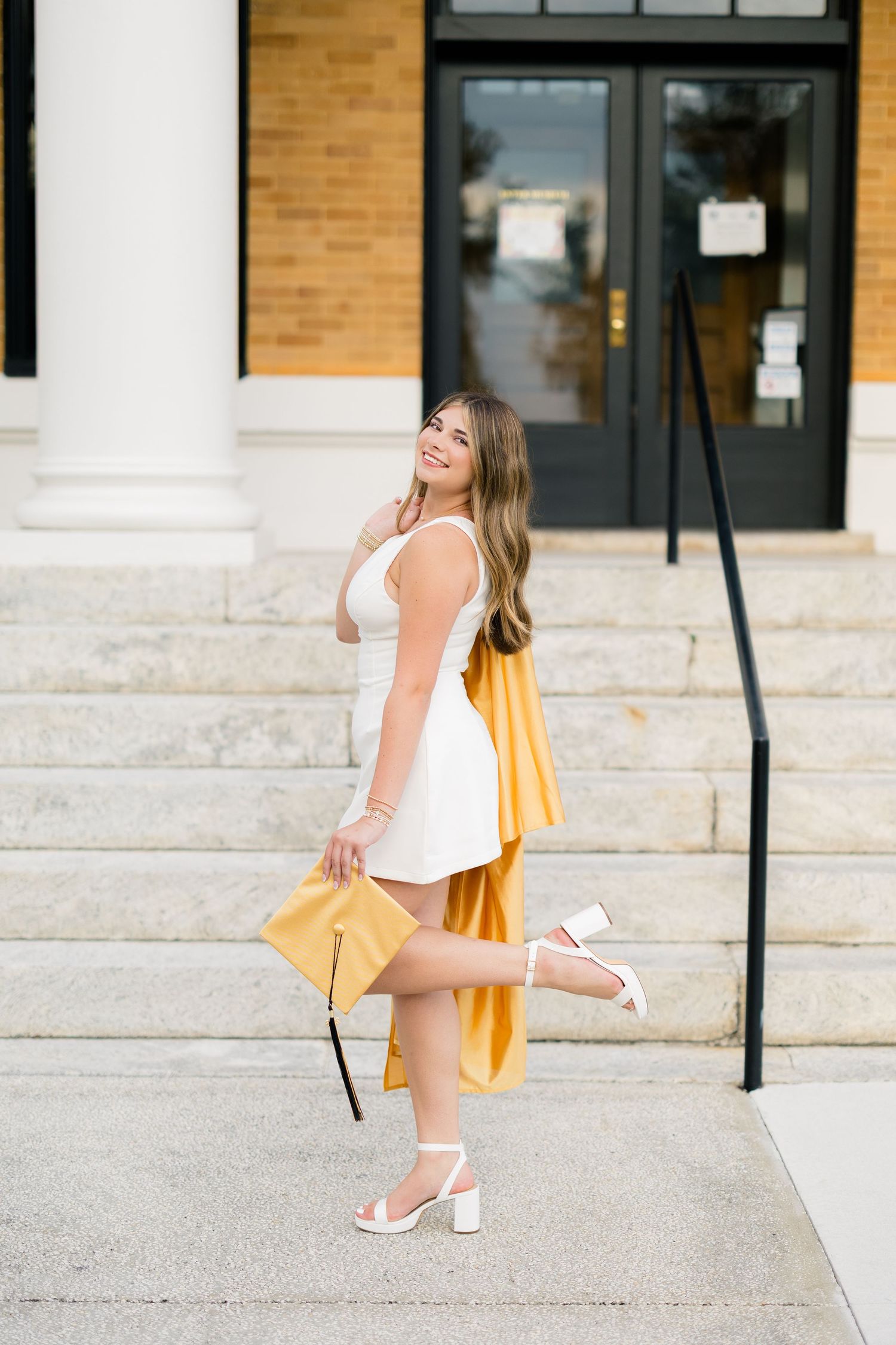 Young woman in white dress and gold graduation gown poses on steps holding cap, celebrating graduation day.
