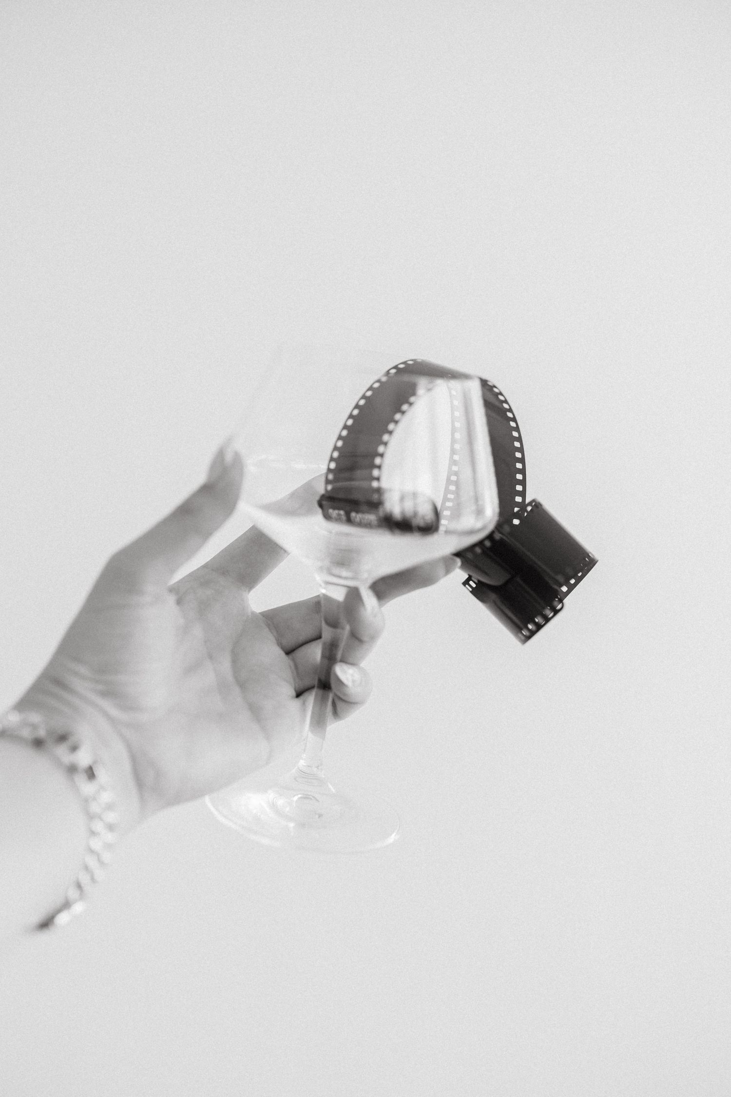 Black and white photo of a hand holding a wine glass with a strip of 35mm film curled inside it.