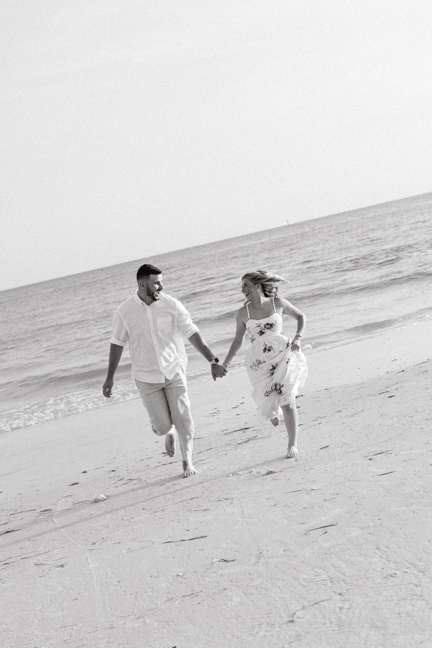 Black and white photo of a happy couple holding hands and running along a sandy beach with ocean waves in the background.