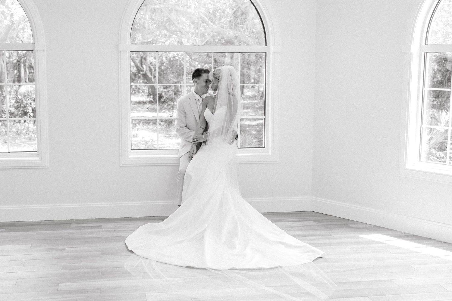 Black and white photo of a bride and groom sharing a kiss in a bright, sunlit room with arched windows.