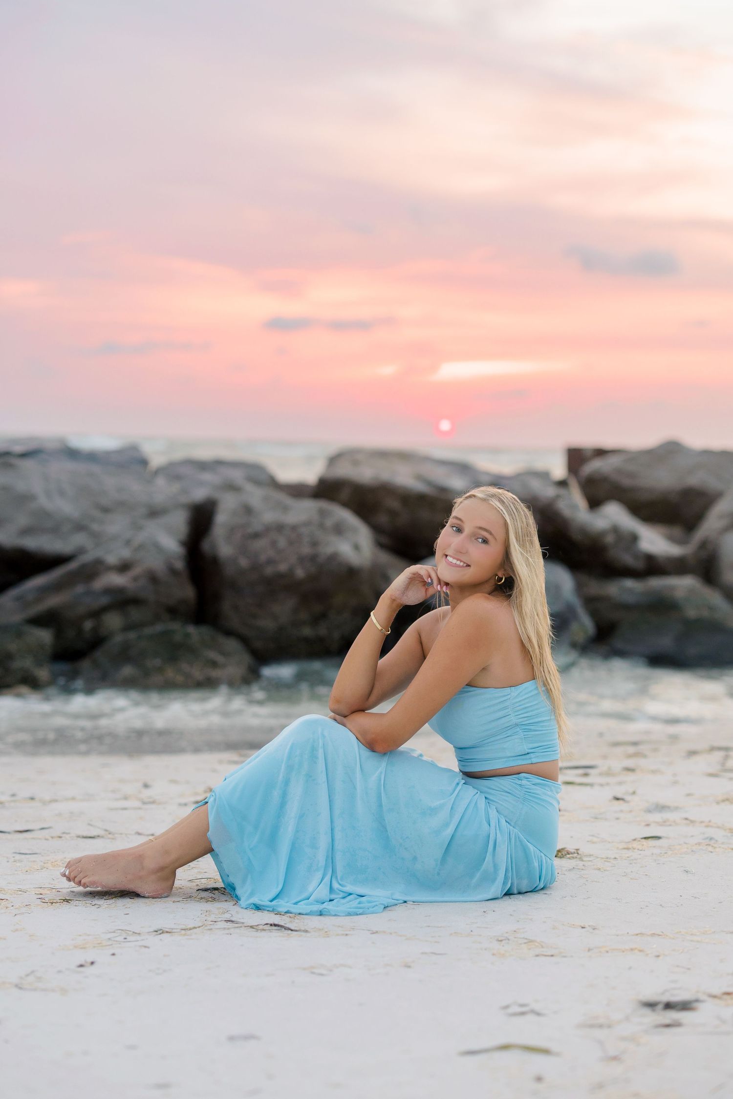 Smiling blonde woman in light blue dress sitting on sandy beach at sunset with rocky jetty backdrop.