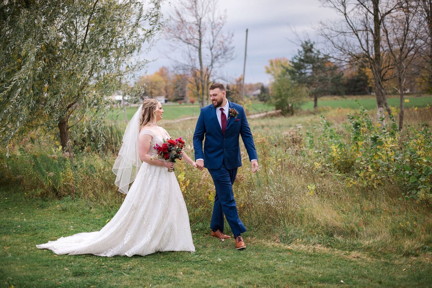 A couple in wedding attire stands together in an autumn field with golden grass and trees in the background.
