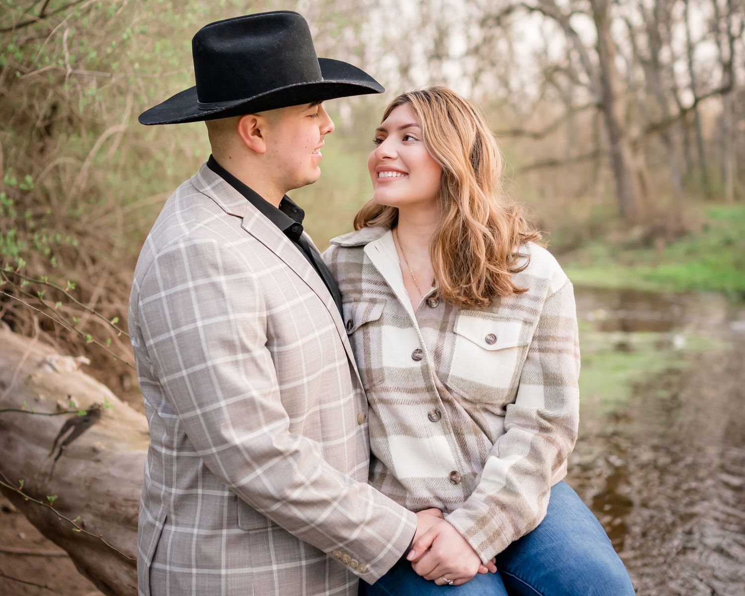A couple in western attire sits together near a creek on a rustic outdoor photo shoot.