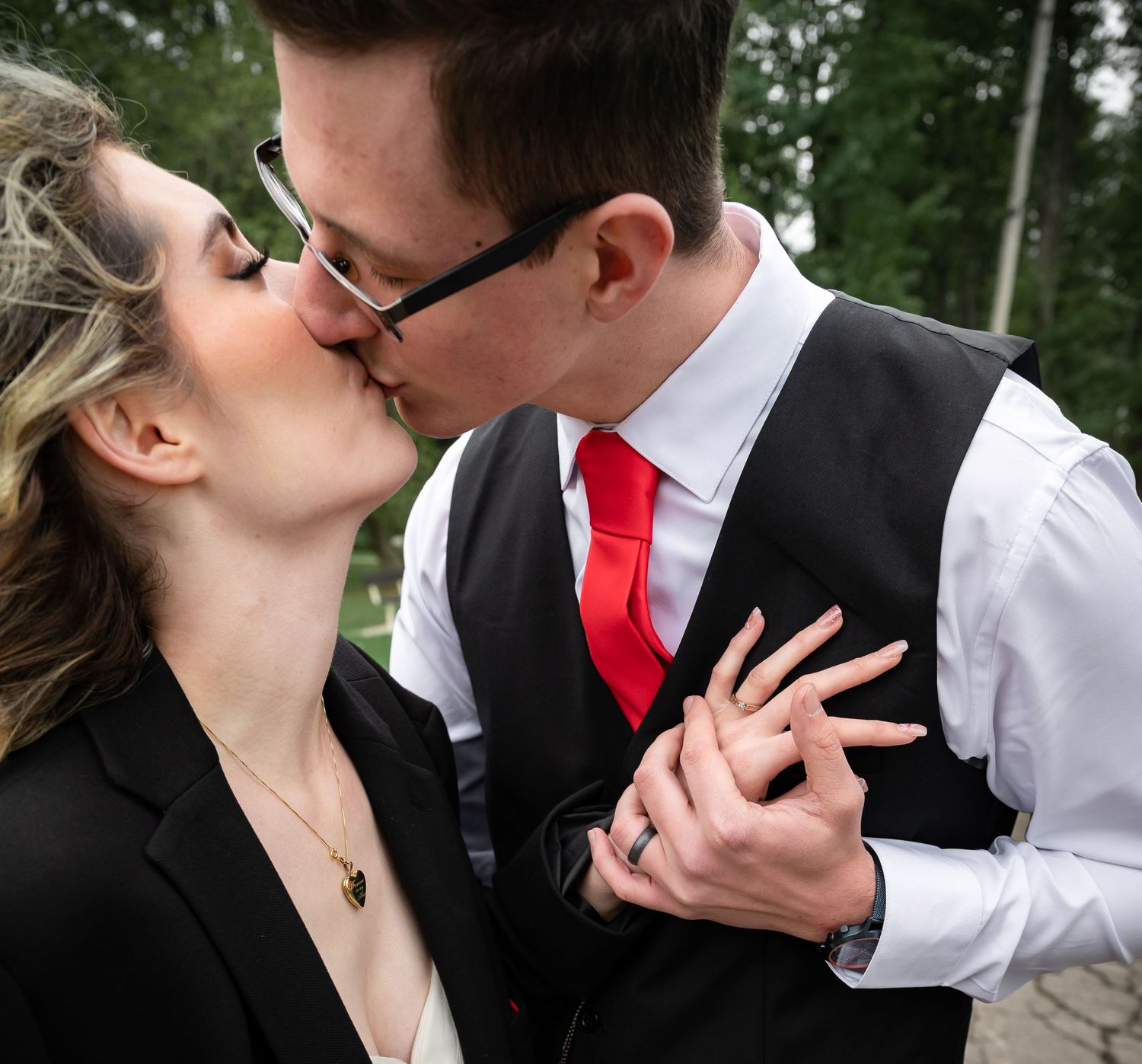 A romantic kiss between a couple, with the groom wearing glasses and a red tie against a natural outdoor background.